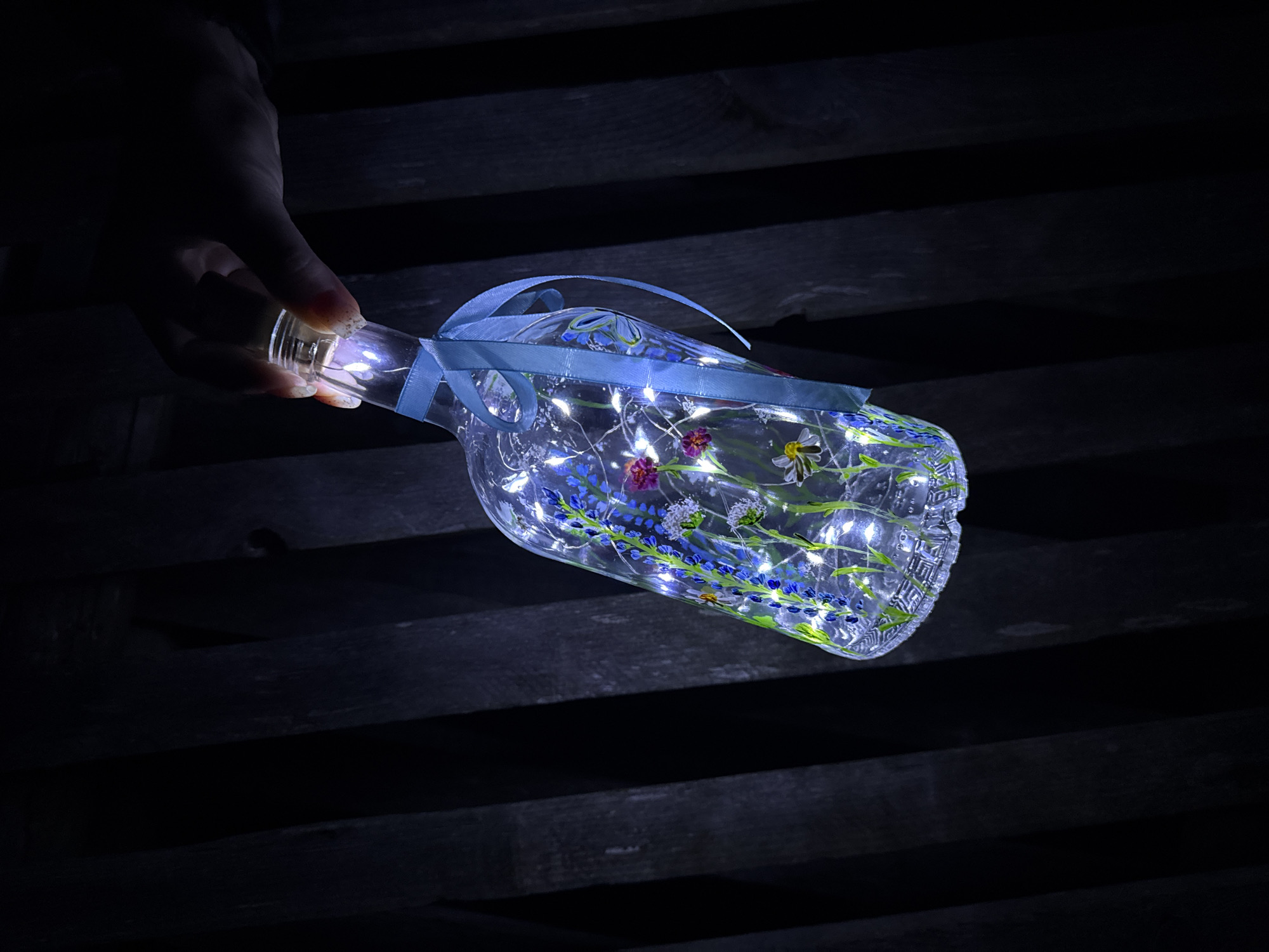 A hand holds a clear glass bottle filled with string lights and decorated with painted flowers and a light blue ribbon. The bottle is positioned over a dark, wooden surface made of parallel planks. The scene is dimly lit, with the bottle and the hand illuminated by the string lights inside it. The location, Вассята, Russia, is not visibly discernible in the image.