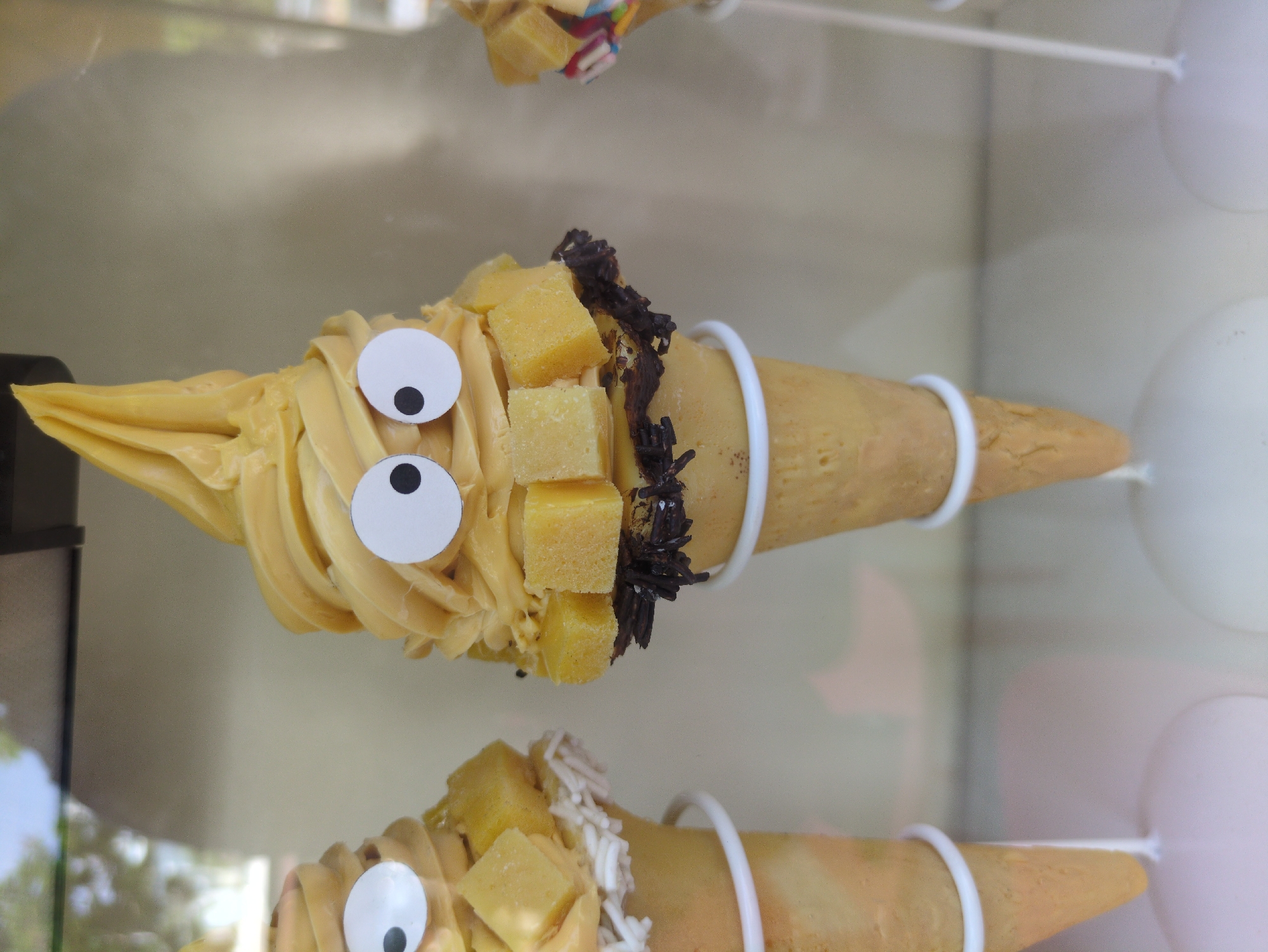 The image depicts two ice cream cones displayed in a glass case. Both cones are filled with yellow ice cream, topped with yellow cubes and adorned with googly eyes. The upper cone also has dark sprinkles around the topping. The cones are held by white rings. The background is blurry, but suggests an indoor setting, potentially a shop. The location is Phú Quốc, Vietnam.