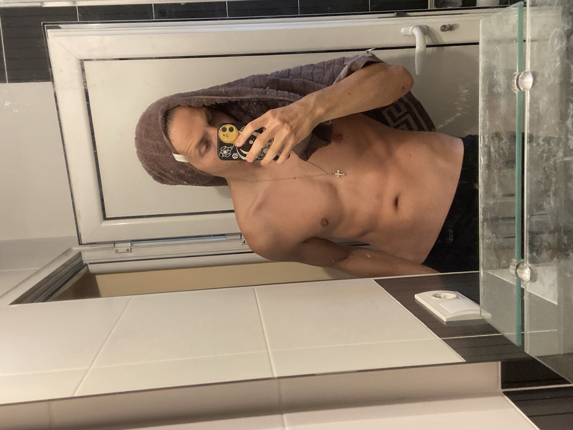 A selfie depicts a shirtless individual with a towel on their head, holding a phone with a patterned case. They wear a necklace with a cross pendant. The setting appears to be a bathroom with black and white tiled walls and a glass shower door. The image was not taken in Šušanj, Montenegro.