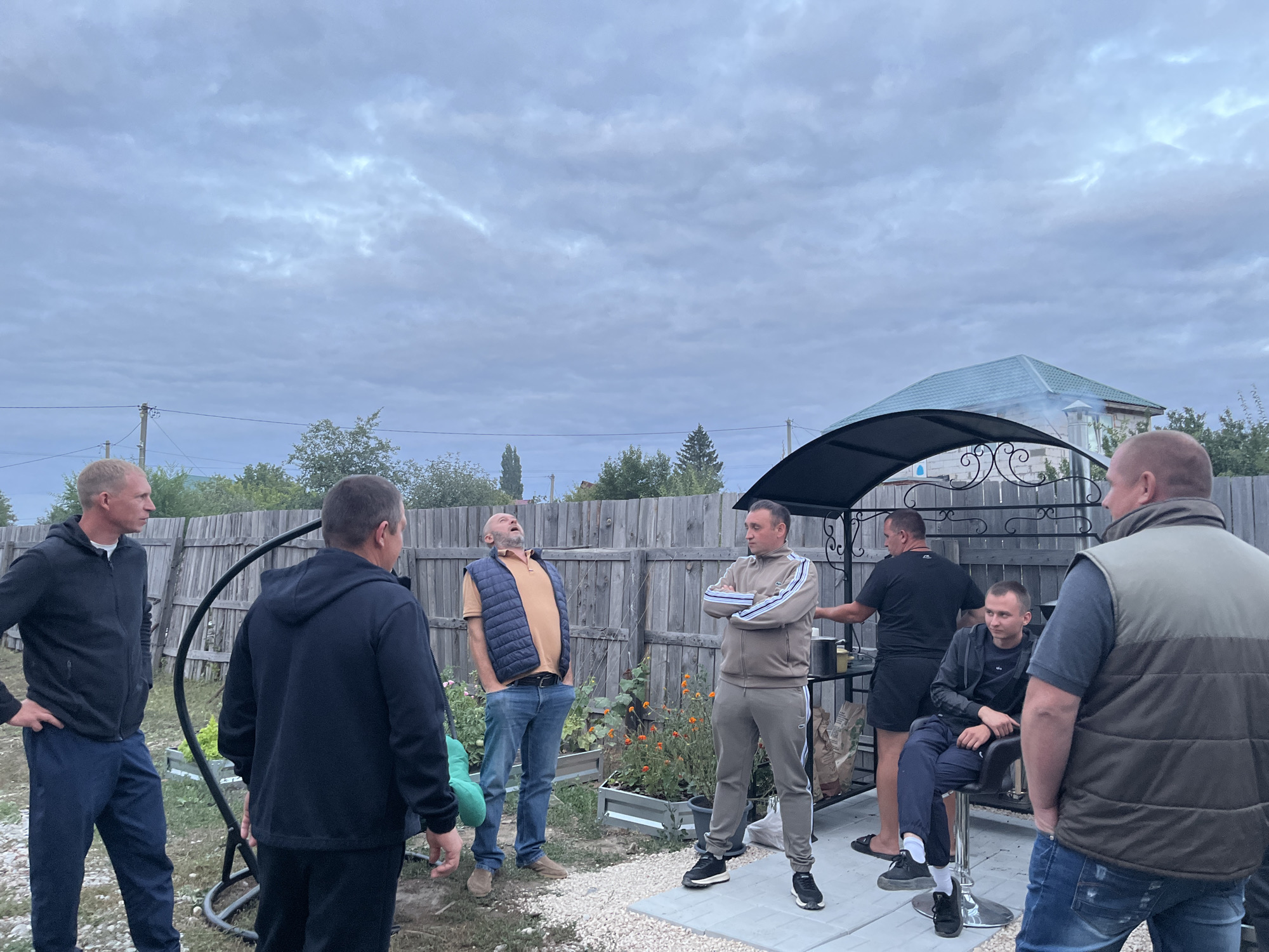 A group of seven men are gathered outdoors in what appears to be a backyard or patio area. The sky is overcast with a cloudy, pale blue hue. In the background, there is a wooden fence, some greenery, and a building with a green roof, possibly a house or shed. A black, ornate gazebo with a dark roof is situated in the middle ground.
The men are casually dressed. Three are standing on the left side of the frame, one in a black hoodie and blue pants, another in a black hoodie, and a third in a tan vest over a brown shirt and blue jeans, with his head tilted back as if laughing or expressing surprise. In the center, a man in a beige tracksuit with blue and white stripes stands with his arms crossed. To his right, a man in a black t-shirt and black shorts stands near the gazebo. On the right, a man sits on a stool wearing a dark jacket over a black shirt and blue pants. The seventh man, on the far right, is partially visible, wearing a dark vest over a blue shirt and blue jeans.
The ground is covered with small white pebbles and some paving stones. There are raised garden beds with some orange flowers visible behind the men on the left. A swing set is also present to the left of the group. The overall impression is of a casual outdoor gathering, possibly a barbecue or social event. The specific location in Saratov, Russia, cannot be determined from the image alone.