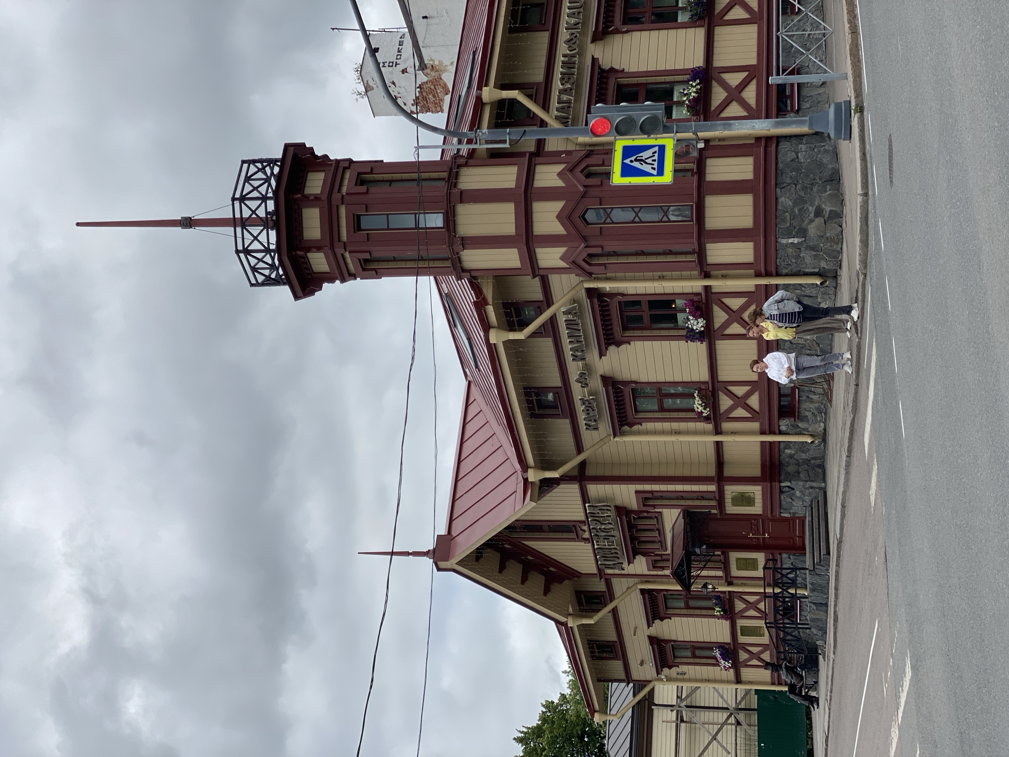 The image captures a street view of the historic "Dom Pervoy Svetopisi" (House of the First Painting) building in Sortavala, Russia. The building is distinguished by its red-and-yellow wooden architecture, featuring a prominent tower with a spire. The facade displays signage in Cyrillic, indicating "Dom Pervoy Svetopisi," "Cafe," and "Shop." Several people are visible: one person is seated on a bench near the entrance, and three individuals are standing on the sidewalk near a pedestrian crossing and traffic light. The sky is overcast, suggesting a cloudy day. The scene includes a traffic light, a pedestrian crossing sign, and a street intersection, indicating an urban setting.