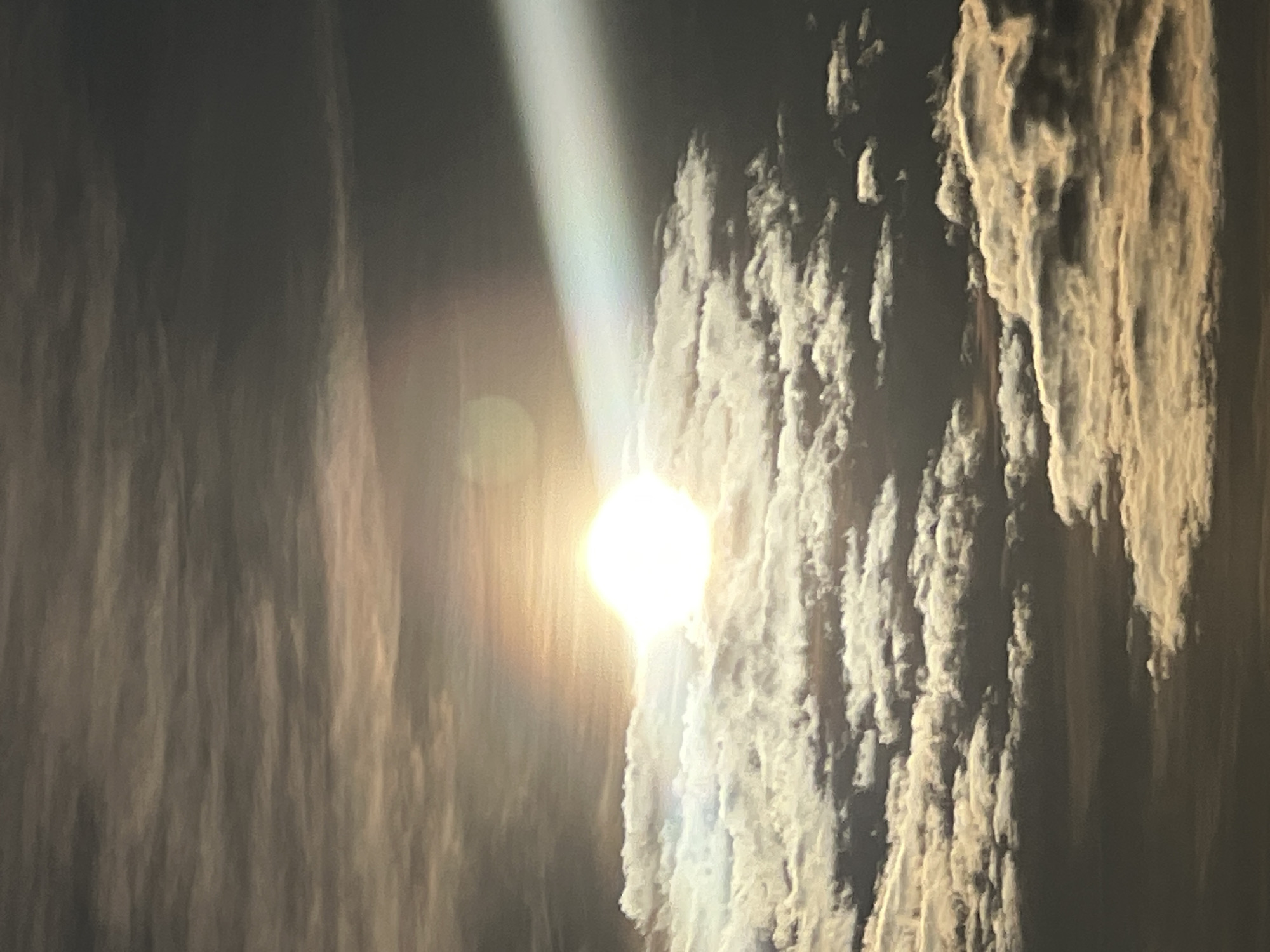 The image shows a daytime sky with the sun shining brightly through a break in the clouds. The clouds are heavily textured and contrast sharply with the dark sky. A single beam of light shines from the sun. There is no evidence to suggest the location is Riga, Latvia.