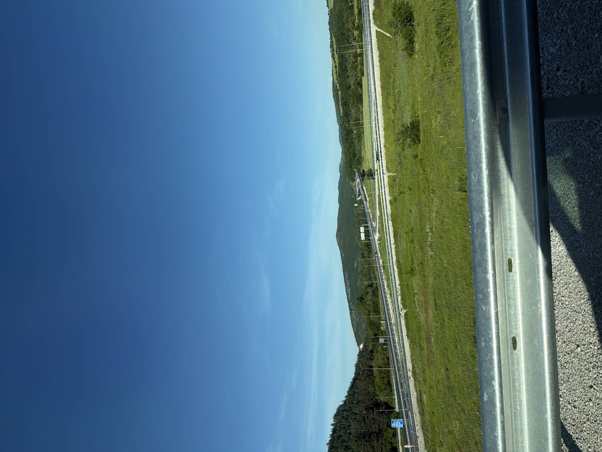 View from a height, possibly a bridge or elevated platform, overlooking the A2 highway in Brestnitsa, Bulgaria. The sky is predominantly clear blue. A metal guardrail is visible in the right foreground. The highway runs parallel to the horizon, flanked by green fields and forested areas. A few cars are visible on the highway. Signage and lighting poles are along the highway.