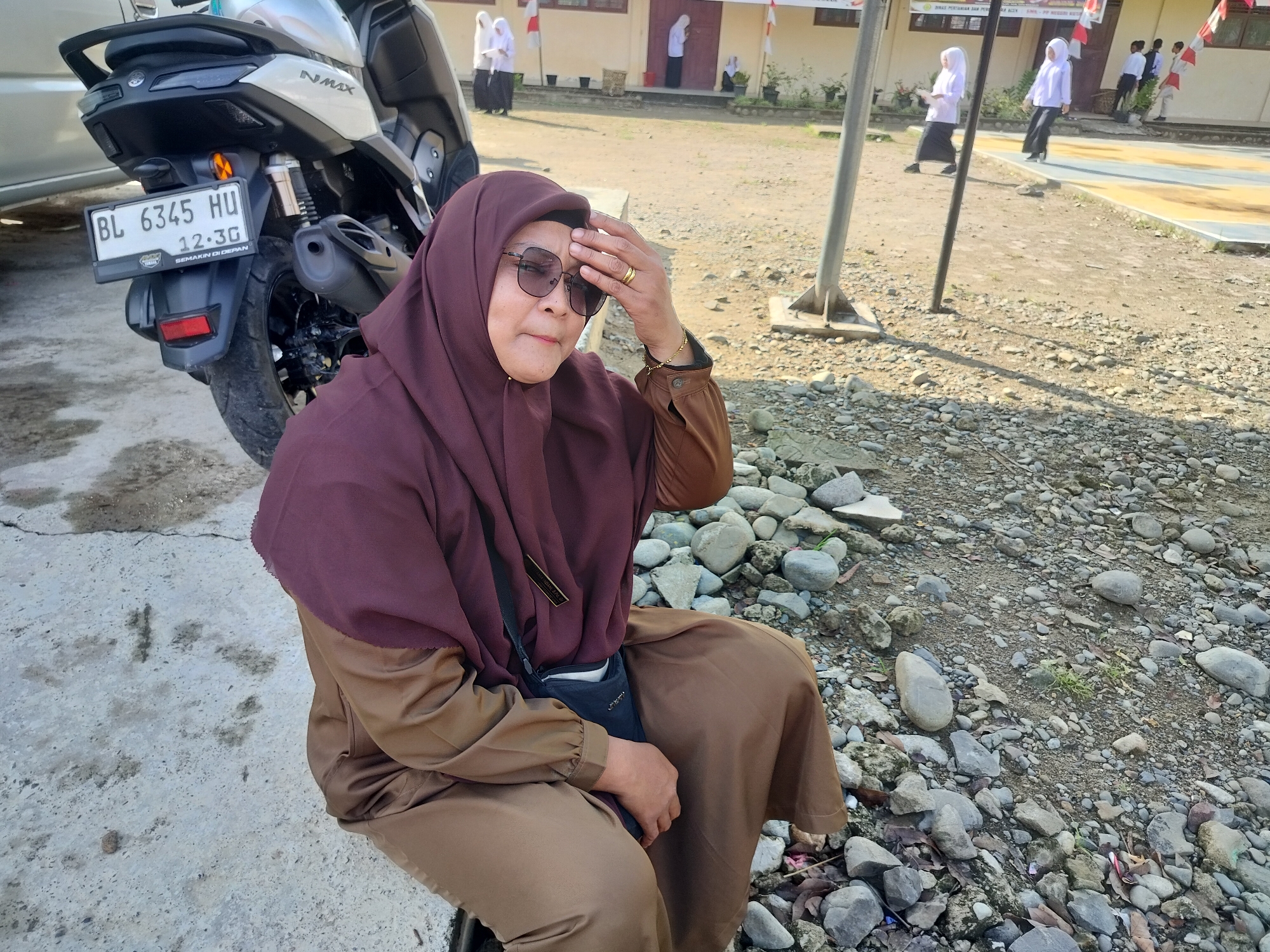 An outdoor, medium shot shows a woman sitting on a concrete curb next to a motorcycle. The woman, who is wearing a maroon hijab, sunglasses, and a brown outfit, has her right hand on her forehead, as if shielding her eyes from the sun. The motorcycle, a silver Yamaha NMAX, has a license plate that reads "BL 6345 HU". In the background, a building with a red door and windows is visible, and several people, some wearing white hijabs and dark skirts, are walking. The ground is covered in gravel and rocks, and there is a metal flagpole in the middle ground. The scene appears to be in the city of Purwodadi, Indonesia, as suggested by the license plate and the architectural style of the building. The lighting suggests it is a sunny day.