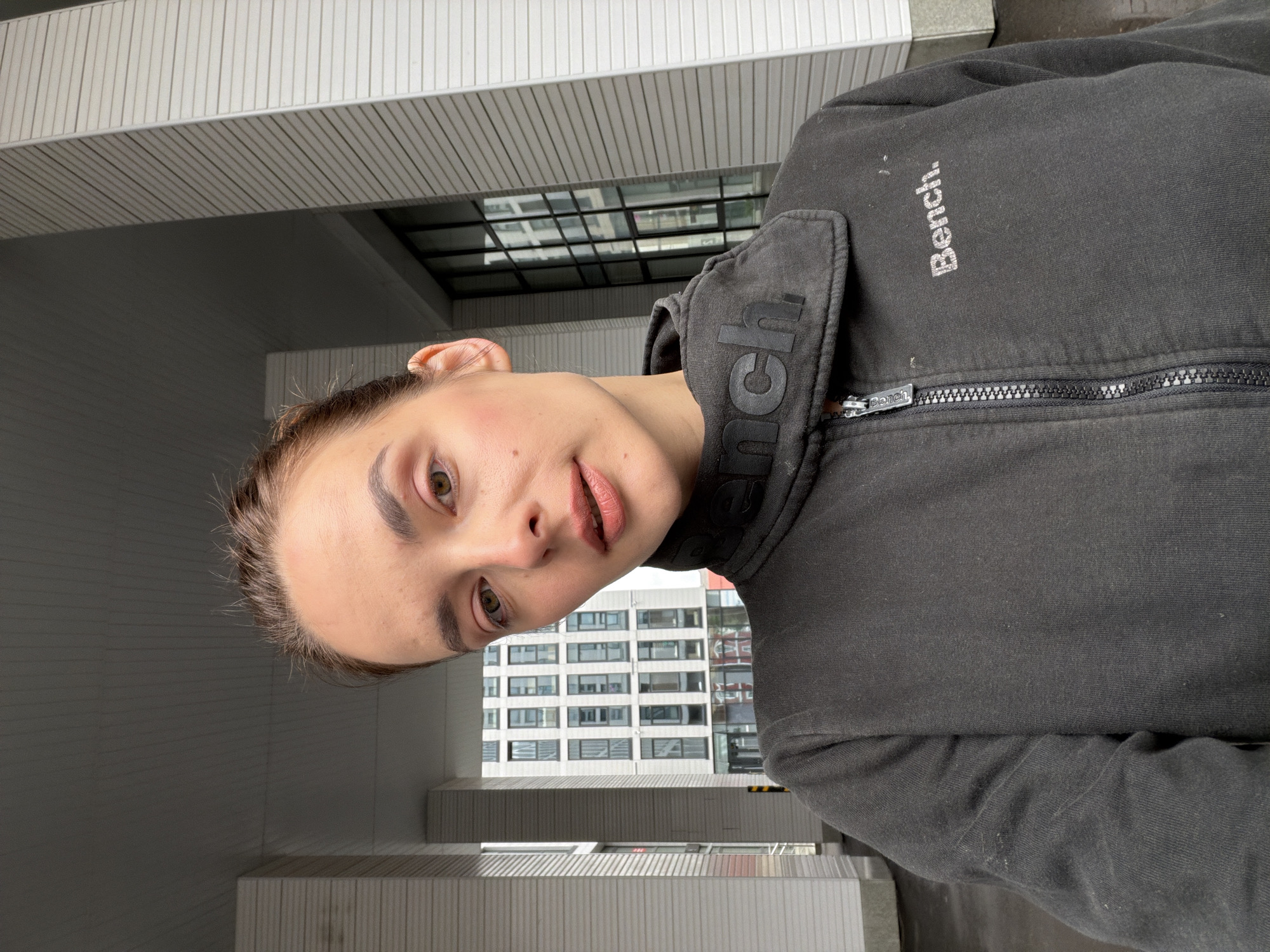 A young woman with brown hair pulled back, wearing a dark gray Bench zip-up jacket, is taking a selfie outdoors. She is looking directly at the camera with a neutral expression, her makeup featuring light eyeshadow and lip gloss. Behind her, there are architectural elements of a modern building, including white columns and ribbed siding. In the background, a multi-story building with numerous windows is visible. The scene appears to be in a city, possibly Ufa, Russia, given the architectural style and the presence of the Bench brand, which is popular in Russia. The overall atmosphere is casual and urban.
