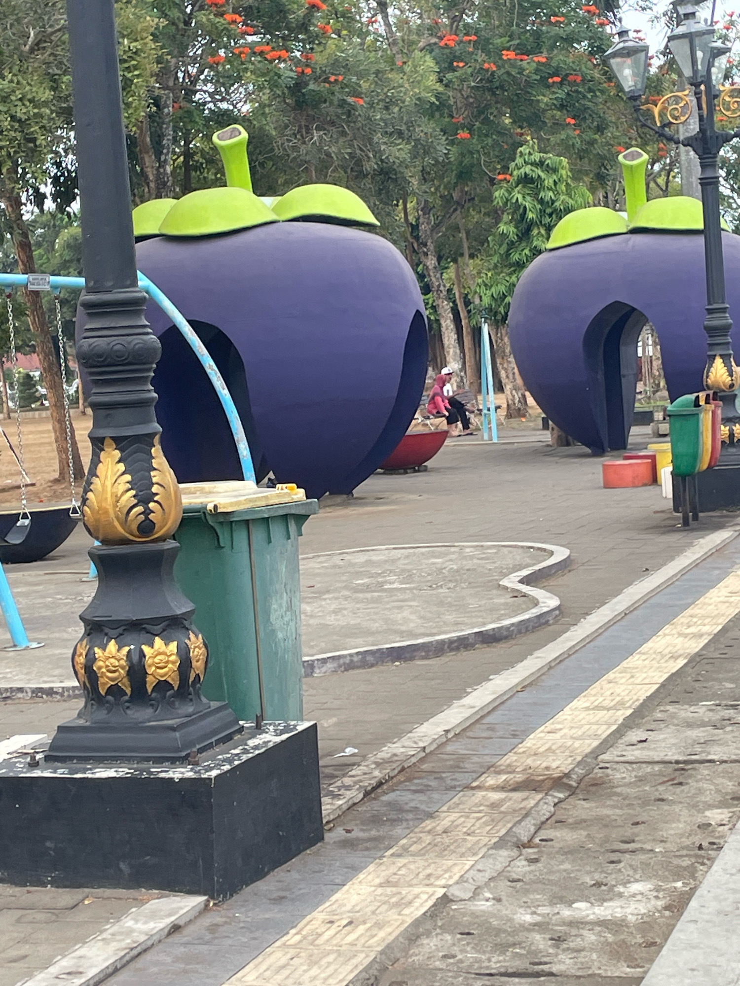 This image depicts an outdoor public space, likely a park or playground, with two large, purple, fruit-shaped structures serving as play areas. These structures resemble mangosteens, with green tops. In the foreground, a black lamppost with ornate gold detailing stands prominently. To the left of the lamppost, a green trash can is visible. Behind the fruit structures, trees with orange blossoms are in bloom. A person wearing a pink headscarf and a pink jacket is seated inside one of the fruit structures, on a red rounded seating element. In the mid-ground, there is a paved pathway with yellow and gray textured tiles. The overall environment suggests a bright, possibly warm day. The context provided indicates the location is the city of Purworejo, Indonesia.