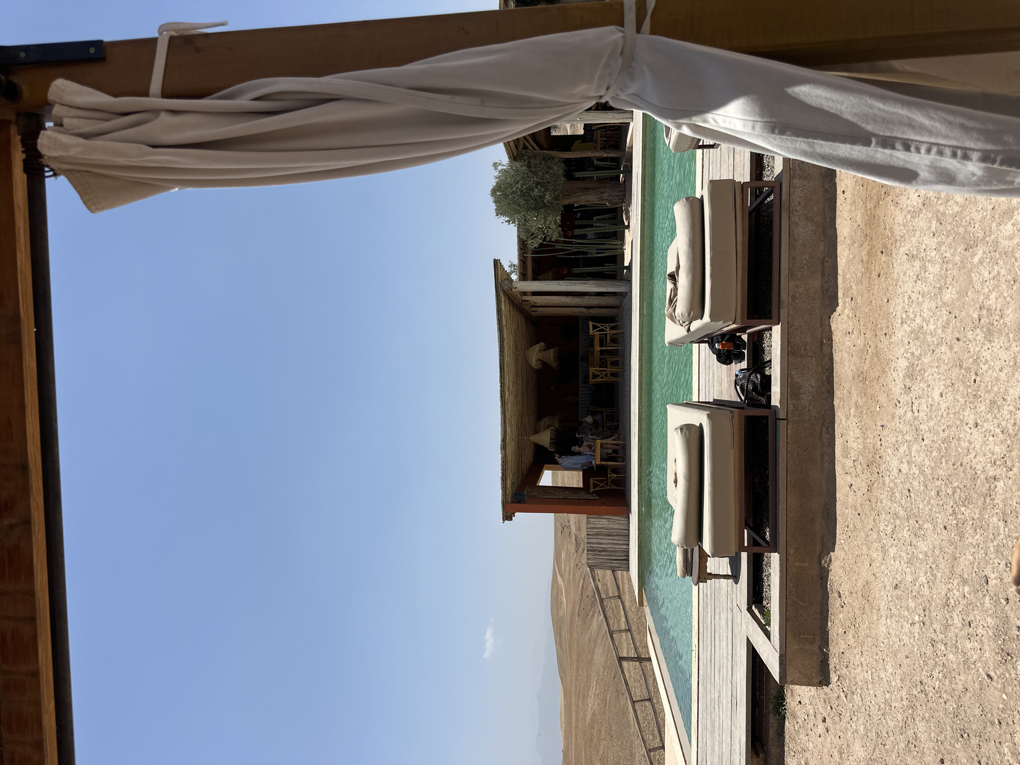 The image depicts an outdoor scene at a location in Sidi Badhaj, Morocco. The foreground shows a lounging area with two cushioned lounge chairs positioned adjacent to a swimming pool. A beige canopy covers the lounging area. In the background, a building with a thatched roof provides a shaded area with tables and chairs where people are seated. Beyond the building, a landscape of arid hills extends into the distance under a clear blue sky. A fence runs along the edge of the pool area, separating it from the hillside.
