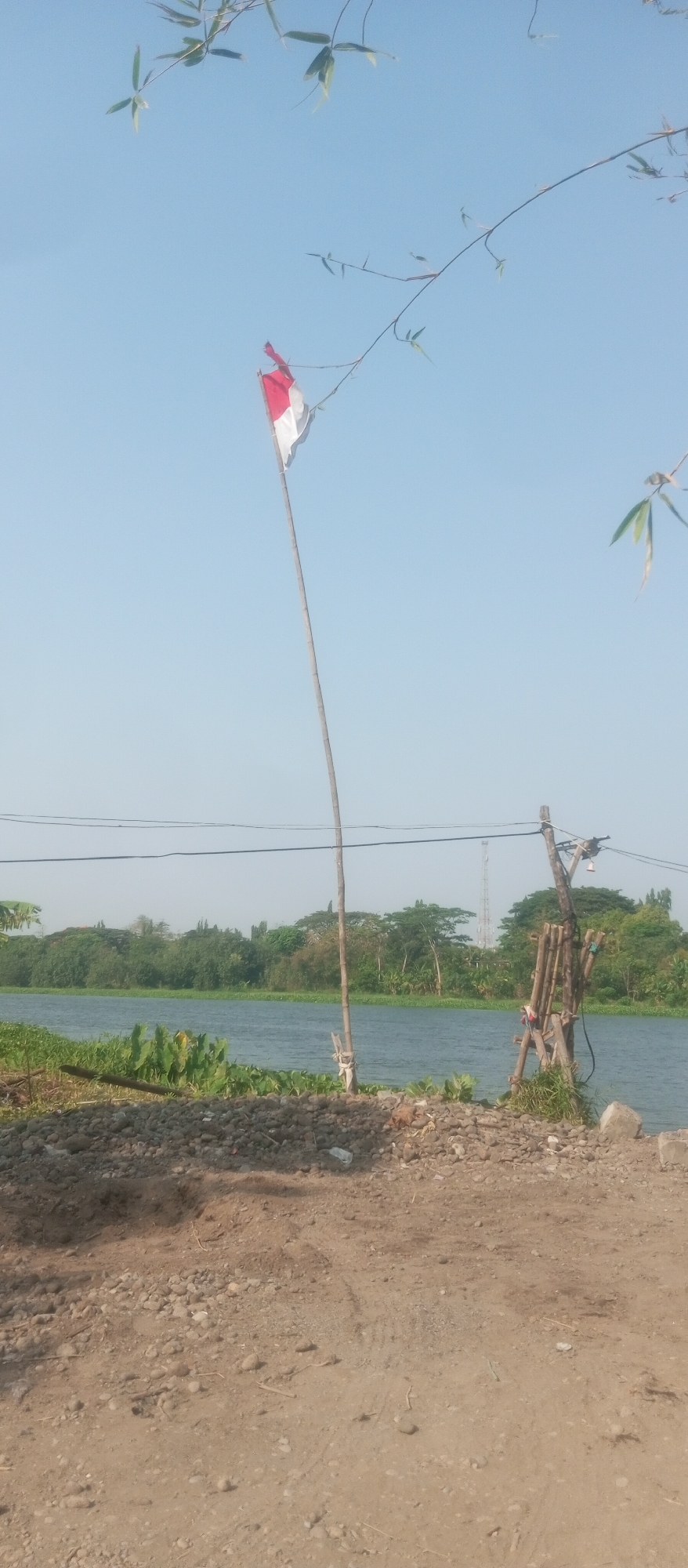 A Dutch flag flies from a tall, thin wooden pole on the bank of a body of water. The flag is red and white. The body of water is lined with green vegetation, and in the background are more trees and some buildings, including a tall radio tower. The foreground is a dirt path with rocks and some debris. The sky is clear and blue.