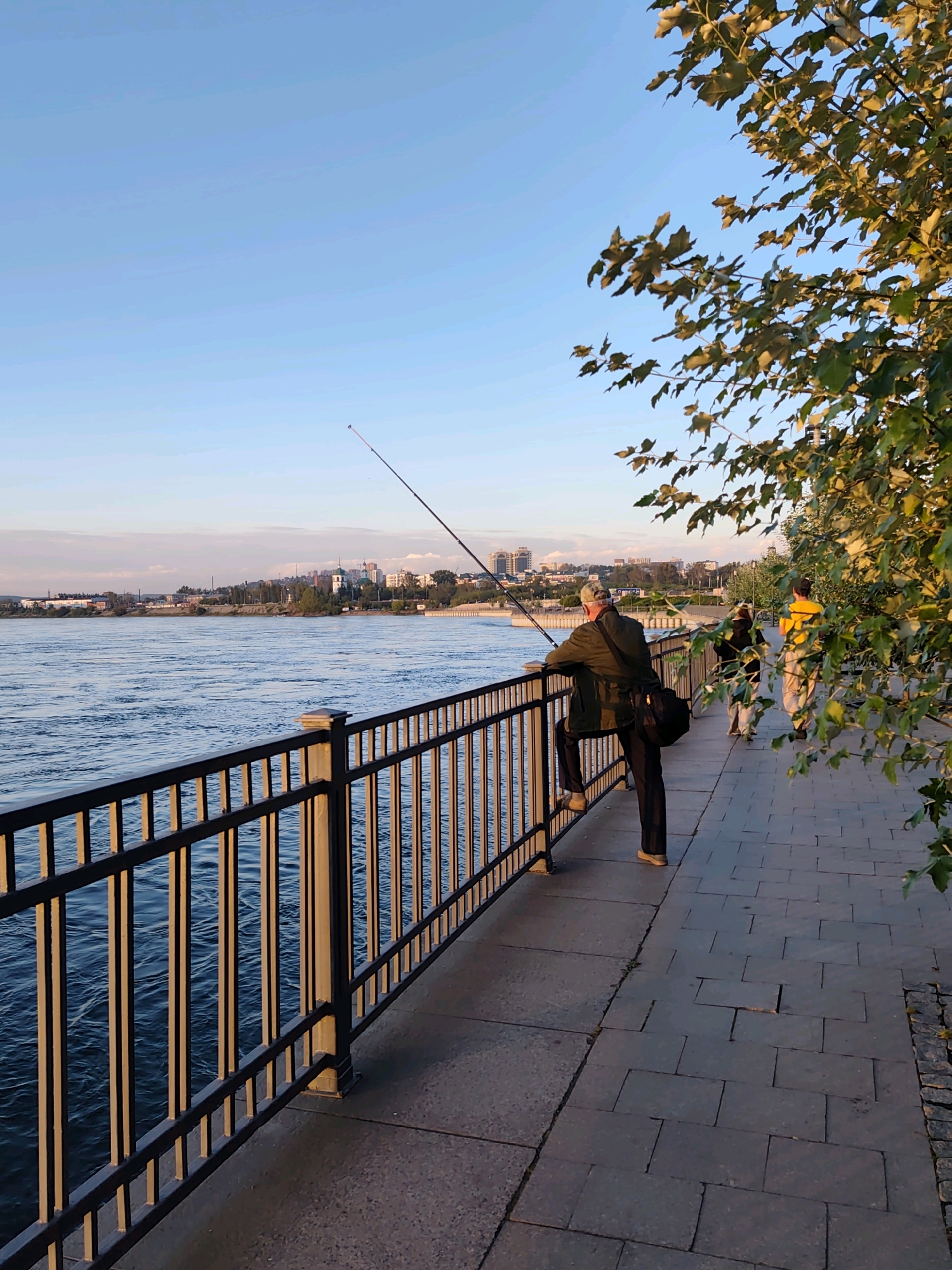 The image depicts a man fishing from a promenade along a wide river, presumably the Angara River in Irkutsk, Russia, given the presence of a city skyline in the background. The man, wearing a cap and a jacket, is leaning over a black metal railing, holding a fishing rod that extends into the water. A dark bag is slung over his shoulder. To his right, two other figures, one wearing a yellow vest and the other in a darker jacket, are walking along the paved promenade. Lush green foliage from a tree overhangs the right side of the scene, catching the golden light of what appears to be late afternoon or early morning sun. The water is a deep blue with ripples, and the city in the distance features a mix of buildings, including what appears to be a domed church. The promenade is paved with dark rectangular stones.