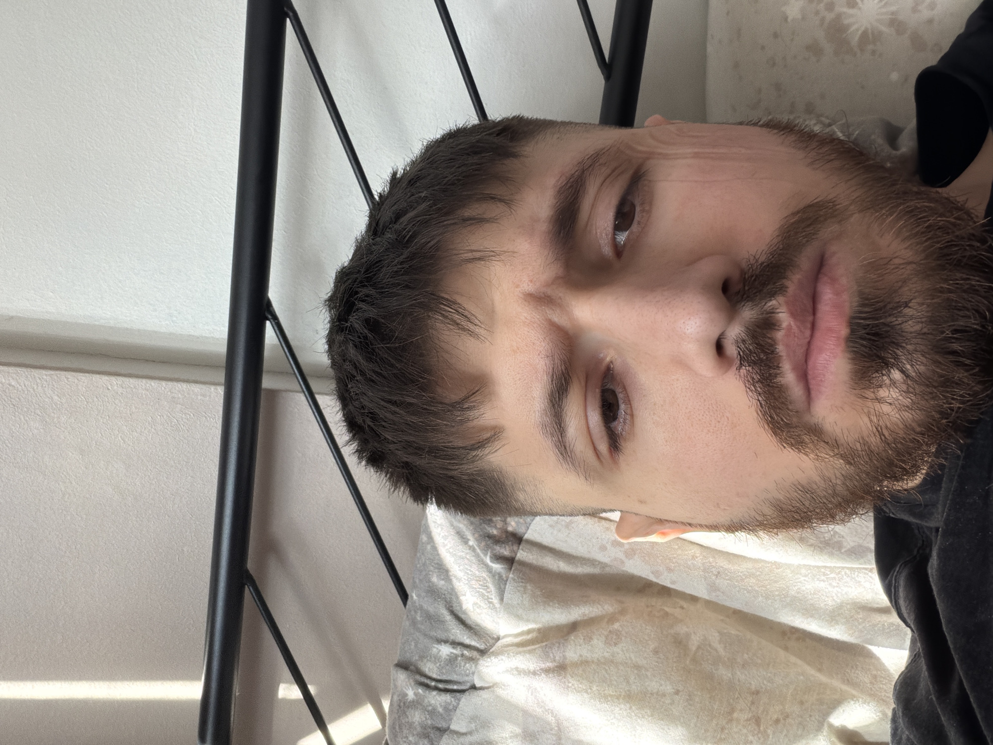 The image depicts a man with brown hair and a beard lying on a bed with a patterned sheet and pillow. He is looking directly at the camera with a neutral expression. A black metal bed frame is visible to the left of the frame. A light-colored wall is behind the bed, partially obscured by the bed frame. The location is not discernible from the provided image.