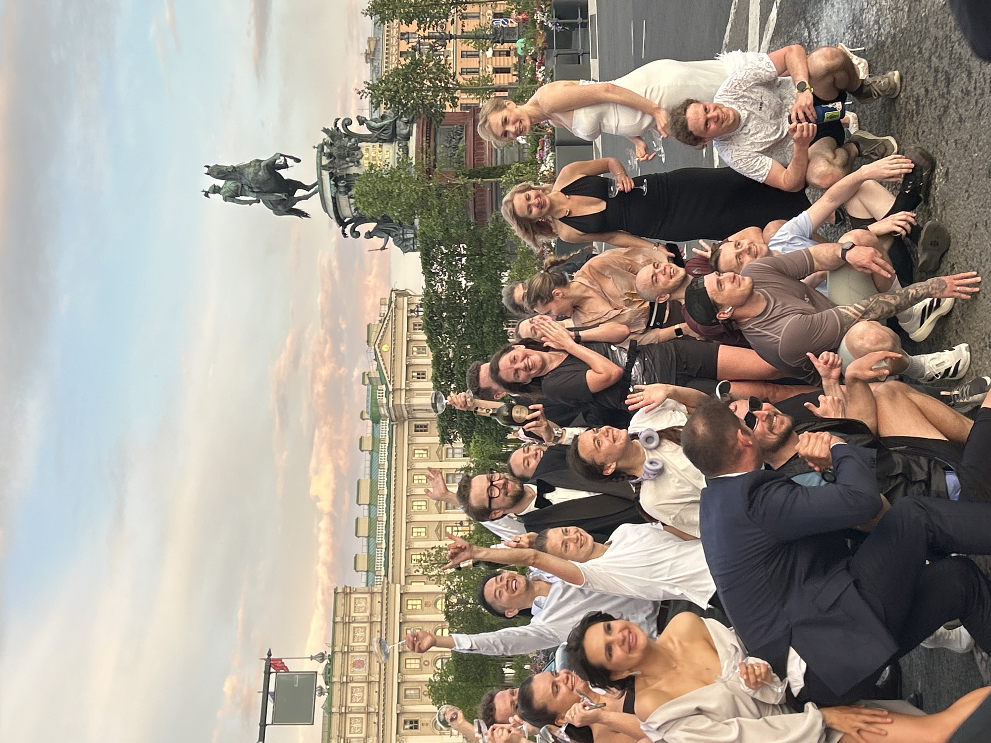 A group of approximately 20 young adults, dressed in a mix of formal and casual attire, pose for a photograph outdoors in front of a large, ornate building with classical architecture. Many are holding champagne glasses, and some are raising them in a toast. The sky above is filled with scattered clouds and a warm, sunset glow. In the background, a large equestrian statue on a pedestal is visible. The ground appears to be paved, possibly a street or plaza. The overall atmosphere suggests a celebratory event.
