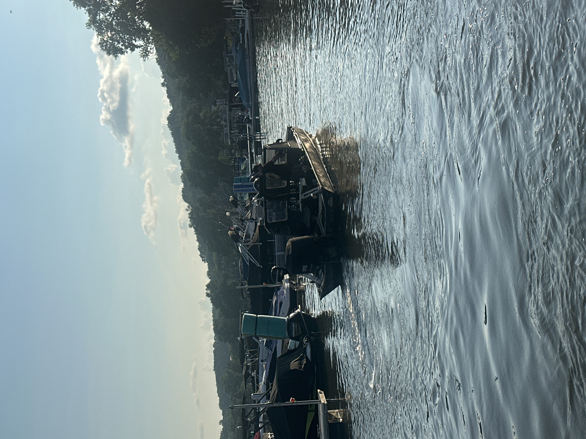 In Algonquin, United States of America, on a bright, clear day, a dark, flat-bottomed boat with multiple engines is moving across the water. The boat has a military or tactical appearance, with dark canvas covers and what appear to be military personnel on board. Several other boats are docked in the background, some covered with tarps. The water is rippled, with sunlight glinting off the surface. Trees and hills are visible in the distance, creating a scenic backdrop for the marina. The sky is a pale blue with a few scattered clouds.