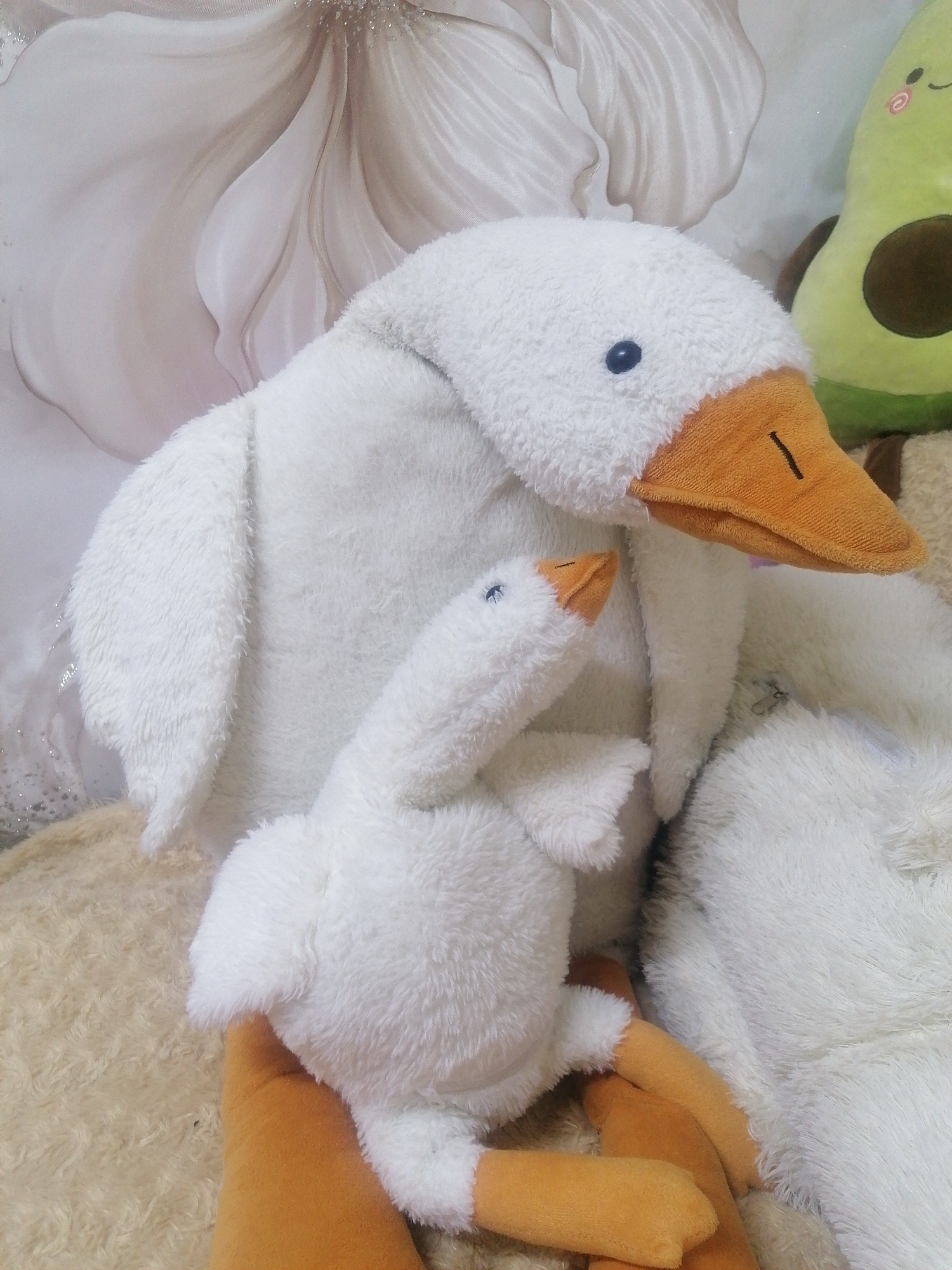 Two white stuffed geese, one significantly larger than the other, are nestled together on a soft, cream-colored rug. The larger goose is prominently featured, its fluffy white body facing towards the viewer, with an orange beak and a single blue eye visible. A smaller goose, also white with an orange beak, is positioned in front of the larger one, its head tilted upwards as if interacting with it. Behind the geese, a portion of a stuffed avocado toy can be seen, adding a touch of vibrant green to the scene. The background features a floral-patterned fabric, likely a wall hanging or tapestry, in muted shades of white and beige, with subtle glitter accents. The lighting appears soft and diffused, suggesting an indoor setting during daylight hours. The overall mood is peaceful and cozy, evoking a sense of comfort and playfulness. There is no visible text in the image.