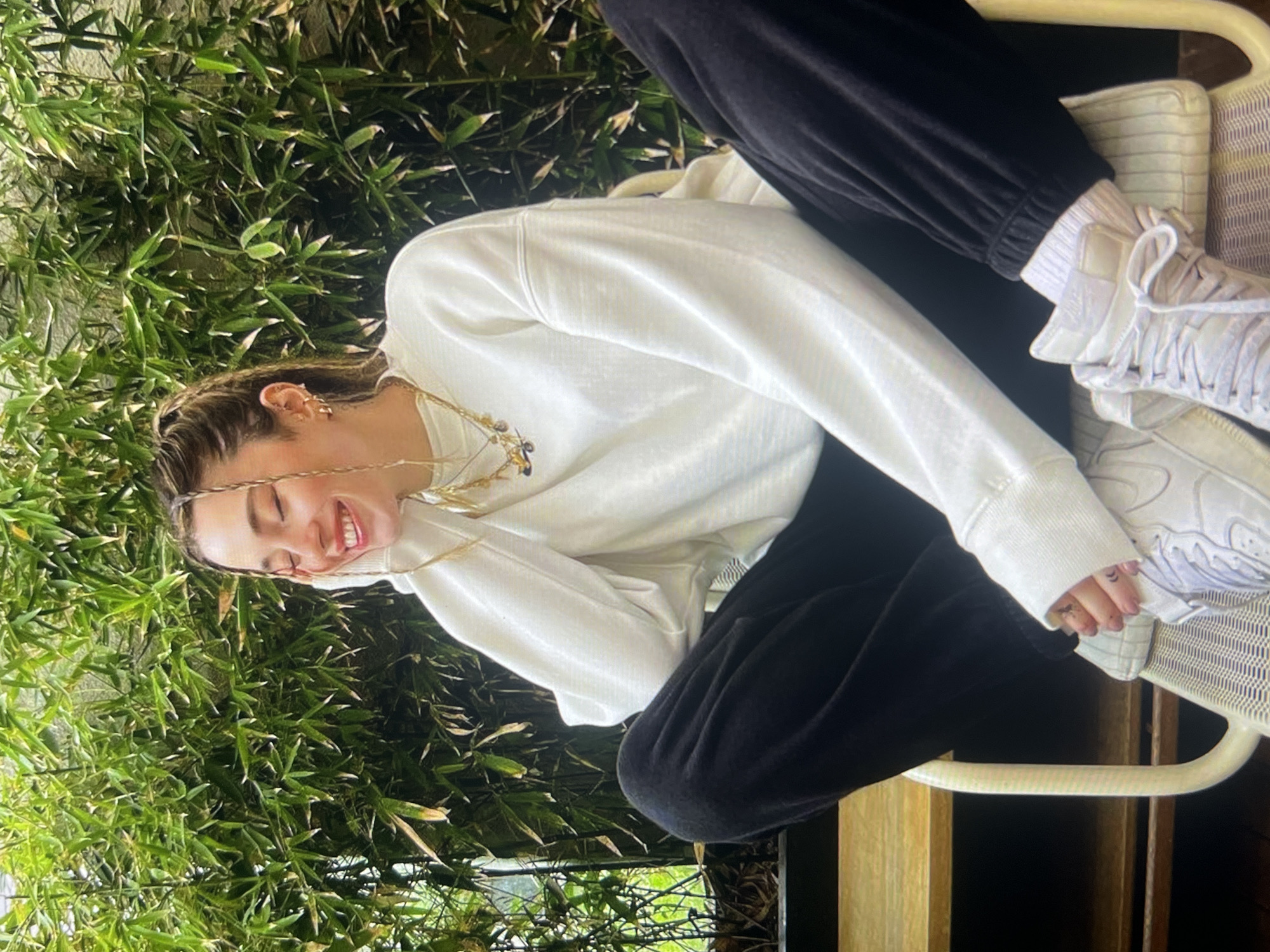 This is a vertical, close-up, outdoor shot of a young woman sitting on a chair. She is wearing a white sweatshirt, black sweatpants, and white sneakers with white socks. Her hair is styled in braids, and she has on a gold necklace. She is smiling with her eyes closed, and her head is tilted back slightly. Her right hand is resting on her head, and her left hand is on her lap. The background is filled with lush green bamboo. The lighting suggests it might be daytime. The overall mood is relaxed and cheerful.