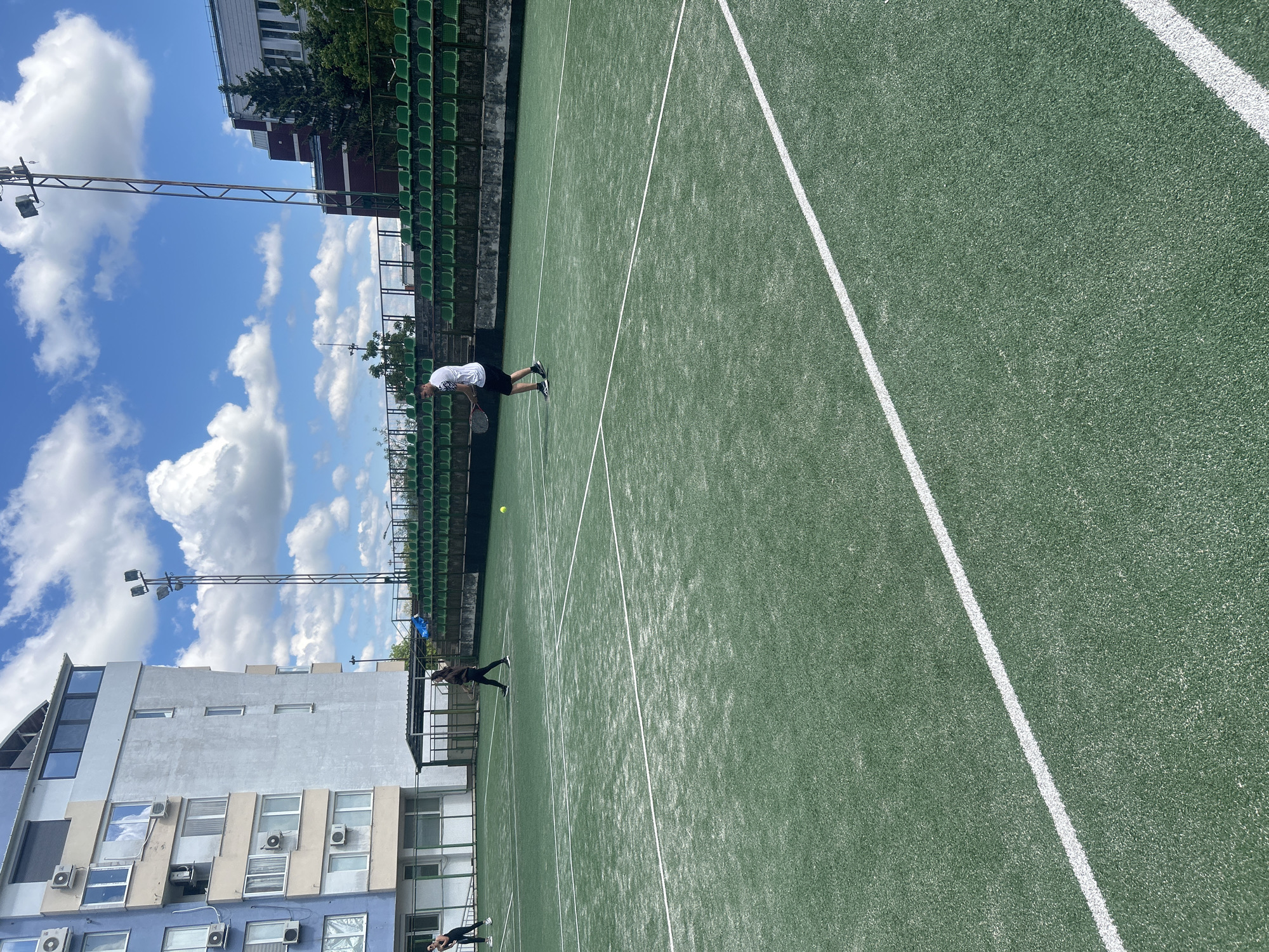 The image depicts a tennis court in what appears to be Veliko Tarnovo, Bulgaria, based on architectural styles and vegetation. The court is green with white lines marking boundaries. Two individuals are present: one is wearing a white shirt and black shorts, appearing to be in the act of swinging a tennis racket. The other individual is wearing dark clothing and is in motion on the court. A tennis ball is visible near the first individual. The court is situated next to a building with multiple windows and air conditioning units. Green seating lines the side of the court. The sky is visible with blue patches and cumulus clouds. Lighting fixtures stand near the court.