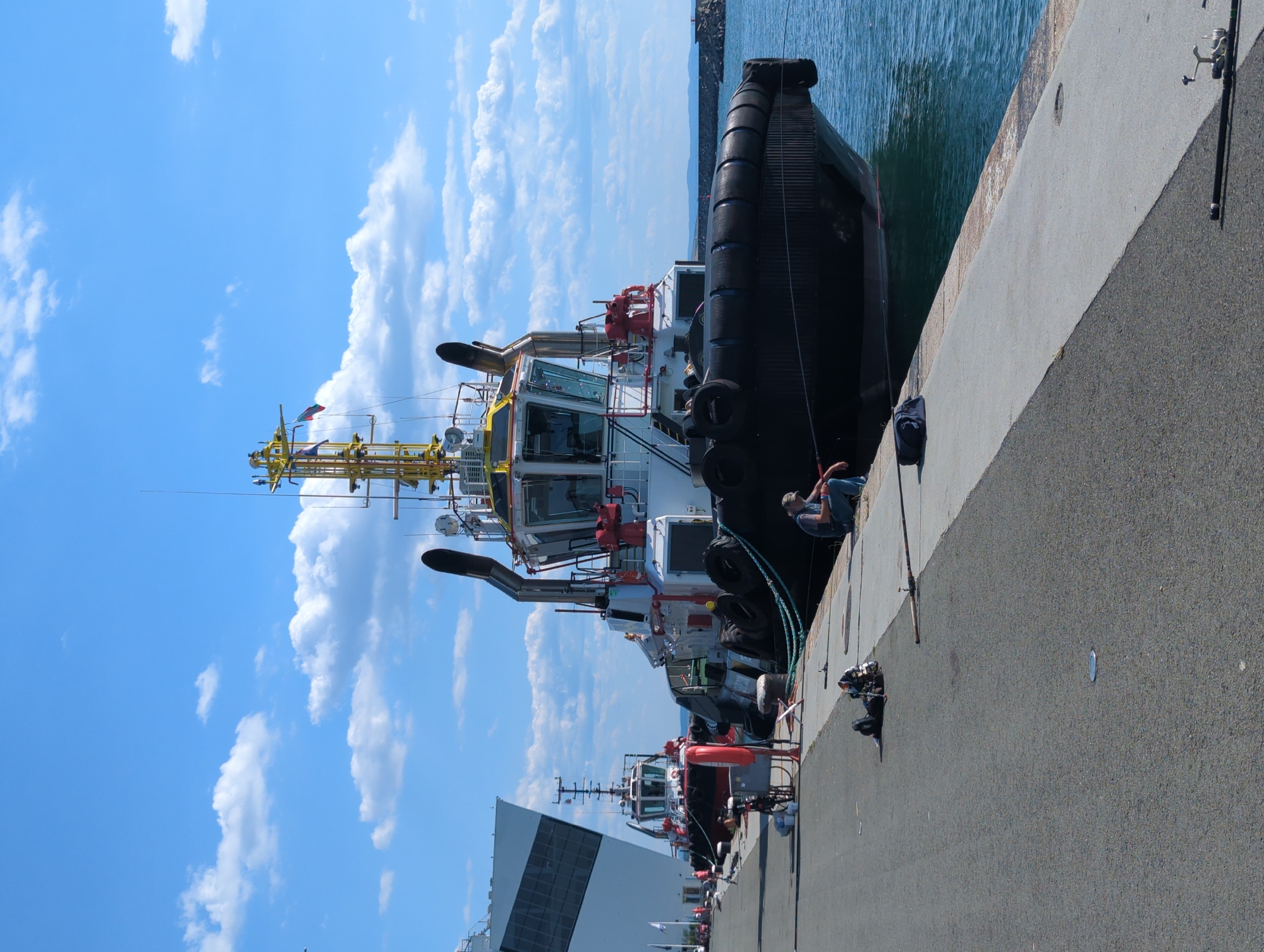 The image depicts a daytime scene at a harbor with a large tugboat docked along a concrete pier. The tugboat is the central focus, featuring a black fendering system, a white and red superstructure, and a yellow crane on top. A person is seated on the edge of the pier, facing away from the camera and towards the water, appearing to be fishing with a fishing rod. In the background, another smaller tugboat is visible, as well as a large, angled, dark-colored structure that may be part of a building or another vessel. The water is calm and blue, and the sky is clear with scattered white clouds. The pier is paved with asphalt and concrete, and various objects are scattered on the ground, including fishing gear and bags. The overall impression is one of a working harbor on a pleasant day.