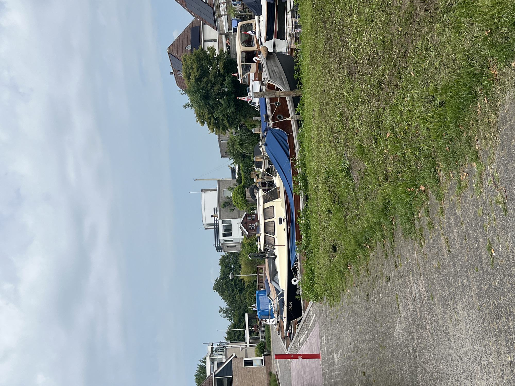 The image depicts a canal in Grou, Netherlands, lined with moored boats. A paved pathway runs along the left side of the frame, bordered by a grassy embankment. Several boats are docked, including a dark-hulled boat with "JIG.CO" written on its side, covered partially by a blue tarp. Another boat with a beige cabin and brown hull is also visible, along with other vessels in the background. Buildings, including houses with solar panels on their roofs, are situated behind the boats and trees. The sky is overcast. There are no people visible in the image.