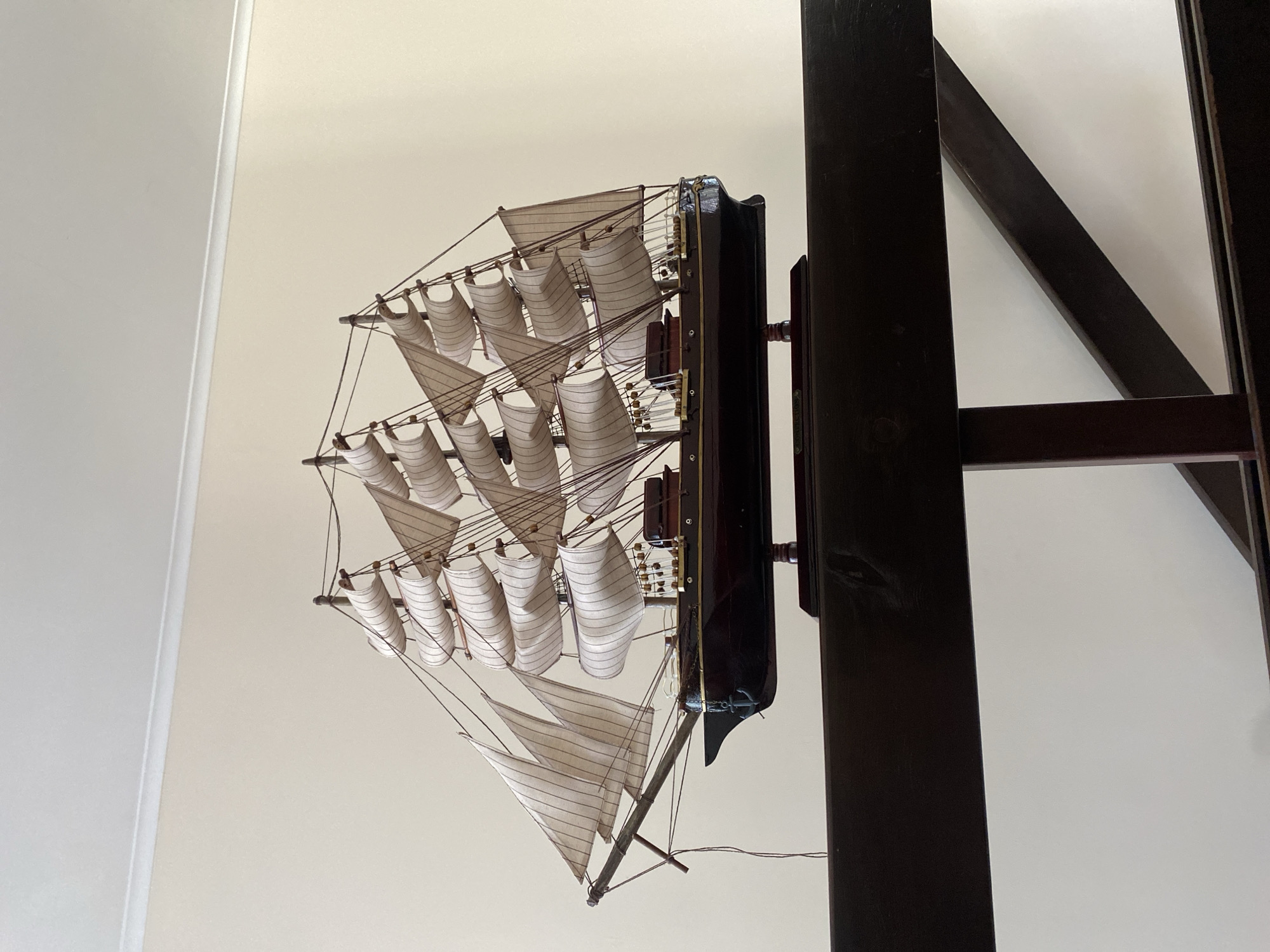 The image shows a model of a sailing ship with white sails, displayed on a dark wooden stand. The ship is positioned in front of a plain, cream-colored wall with white trim. The foreground is dominated by a dark wooden railing, behind which the ship model is visible. The scene appears to be an indoor setting, possibly a home or a decorative display. There is no information or visual evidence to suggest this image is located in the city of Chornomorsk, Ukraine.