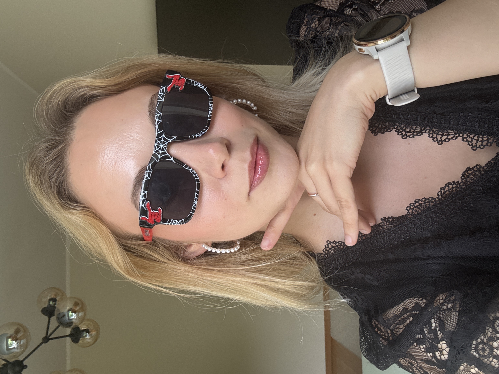 A woman with blond hair wears Spiderman-themed sunglasses and a black lace top. She has pearl earrings and a smartwatch on her left wrist. The background includes a wall, a light fixture with multiple bulbs, and part of a doorway. It is not possible to determine the location within Riga, Latvia.