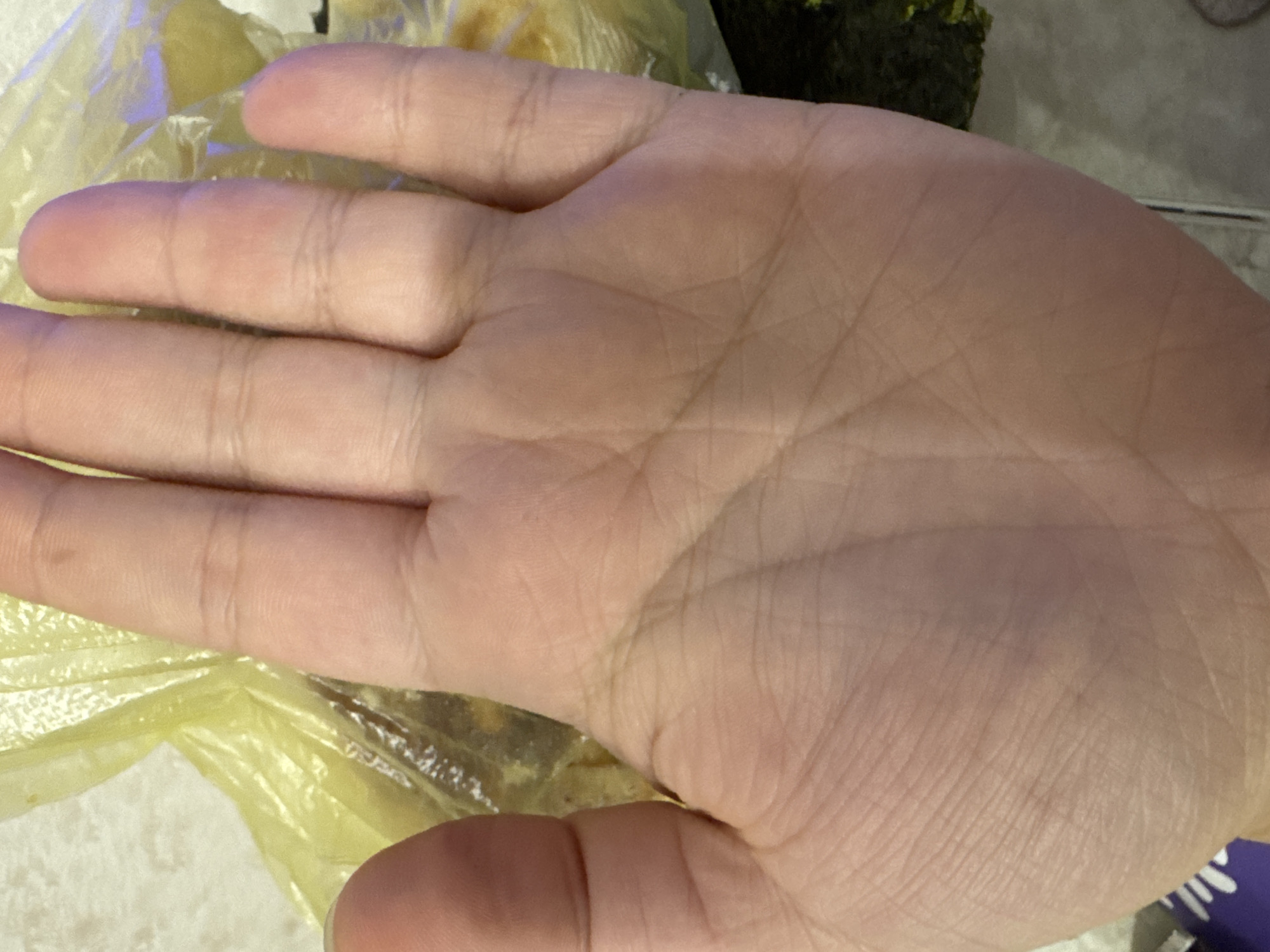 The image shows a close-up view of a person's palm, with fingers extending upwards. The skin is lightly wrinkled, and various palm lines are visible. Behind the palm, a blurred yellow plastic bag is visible, containing what appears to be food items. To the right and bottom of the frame, a dark green, leafy substance, possibly seaweed, is also present. The background is out of focus, but appears to be a textured surface, possibly a countertop or floor. The image does not contain any visual elements that would indicate a specific location in Saratov, Russia.