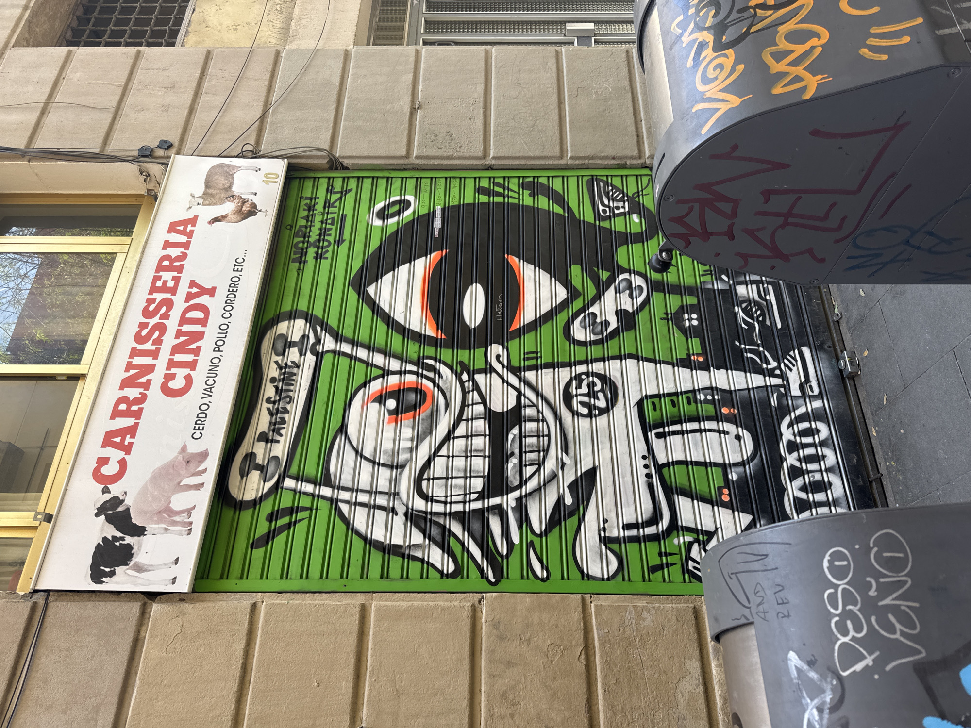 The image shows a storefront in Barcelona, Spain. The roll-down metal gate is painted green and features graffiti-style artwork. The artwork includes abstract figures and an eye. To the left of the gate is a sign for "Carnisseria Cindy," a butcher shop. The sign lists the types of meat sold: pork, beef, chicken, lamb, etc. There is also another graffiti inscription that says "PALESTINE". To the right of the gate are gray cylindrical bollards covered in graffiti. The bollard closer to the viewer has "PESO VENO" spray-painted on it.