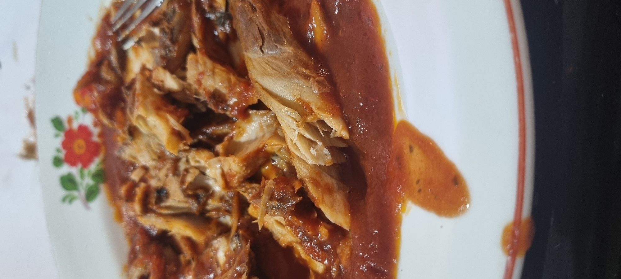 A close-up, slightly overhead shot captures a meal consisting of shredded or flaked meat generously covered in a thick, reddish-brown sauce. The dish is served on a white plate adorned with a floral pattern on the left side, featuring a red flower and green leaves. A silver fork is partially visible in the upper left corner, poised over the food.
The meat displays a tender, fibrous texture with individual flakes evident, suggesting it has been pulled or slow-cooked. The sauce is a deep, earthy red-brown, appearing rich and somewhat chunky, with small dark specks that may be herbs or spices. A lighter, more orange-hued section of sauce is visible on the plate's right, suggesting a slight separation or varied consistency. The plate itself has a thin reddish-orange rim.
The setting appears to be indoors, with a dark, out-of-focus background on the right, likely a table surface. The lighting is somewhat soft, highlighting the textures of the food and the sheen of the sauce. No specific time of day, location, or emotions are discernible, and no text is visible in the image.