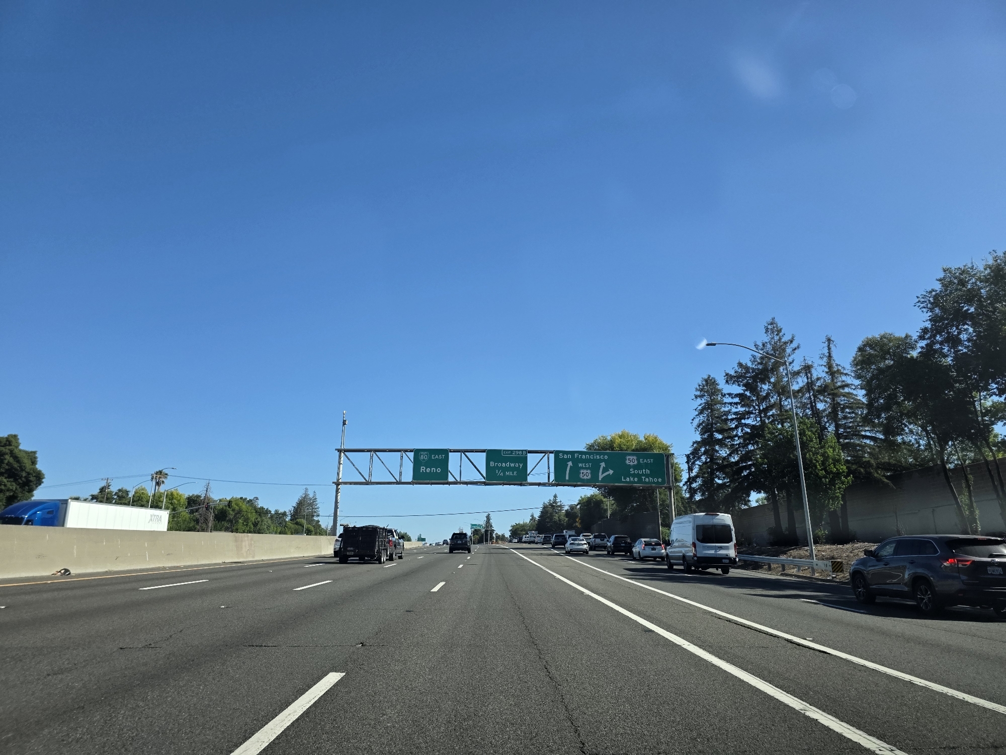 This media file captures a view from a vehicle traveling on a highway in Sacramento, United States of America. The scene is set under a clear, bright blue sky, indicating fair weather and likely daytime. The highway stretches ahead, with multiple lanes of asphalt marked by white lines. Several vehicles are visible on the road, including cars, vans, and trucks, suggesting moderate traffic.
Overhead, a large gantry displays green highway signs. The signs provide directional information, indicating exits and destinations. Text visible on the signs includes "80 EAST Reno," "EXIT 298 B Broadway 1/4 MILE," and "San Francisco WEST" along with "50 EAST South Lake Tahoe." These signs suggest the vehicle is on a route that connects to these major destinations.
On the right side of the highway, trees and a sound barrier line the roadside. On the left, a concrete barrier separates the lanes from what appears to be an embankment or industrial area, with a large white truck visible in the distance. The overall impression is a typical highway scene on a sunny day, with clear visibility and a sense of forward movement. The presence of the highway signs and the general landscape suggest an urban or suburban setting within the United States.