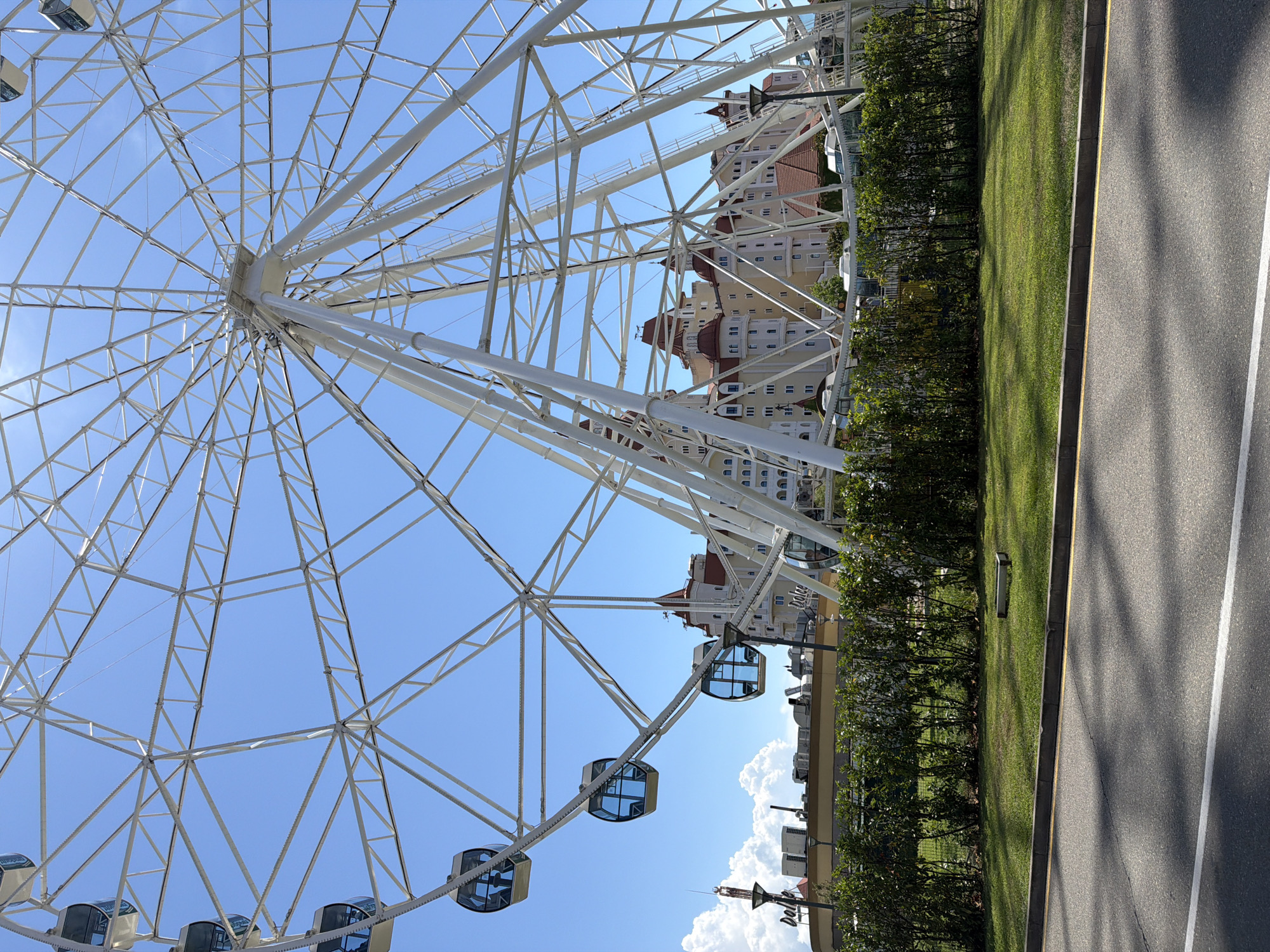 This image captures a close-up view of a Ferris wheel against a clear blue sky. The Ferris wheel is white and features a complex structure of beams and cables. Several enclosed cabins, tinted blue, are visible hanging from the wheel. In the background, a multi-story building with a distinctive red-roofed design and white walls is partially visible. The building appears to be a hotel or resort, consistent with the Olympic Park area in Sochi, Russia, specifically the Sirius Federal Territory. In the foreground, a manicured lawn and a row of green bushes are visible, with a section of asphalt road in the very bottom of the frame. The lighting suggests it is daytime, likely sunny. There are no people clearly identifiable in the image.