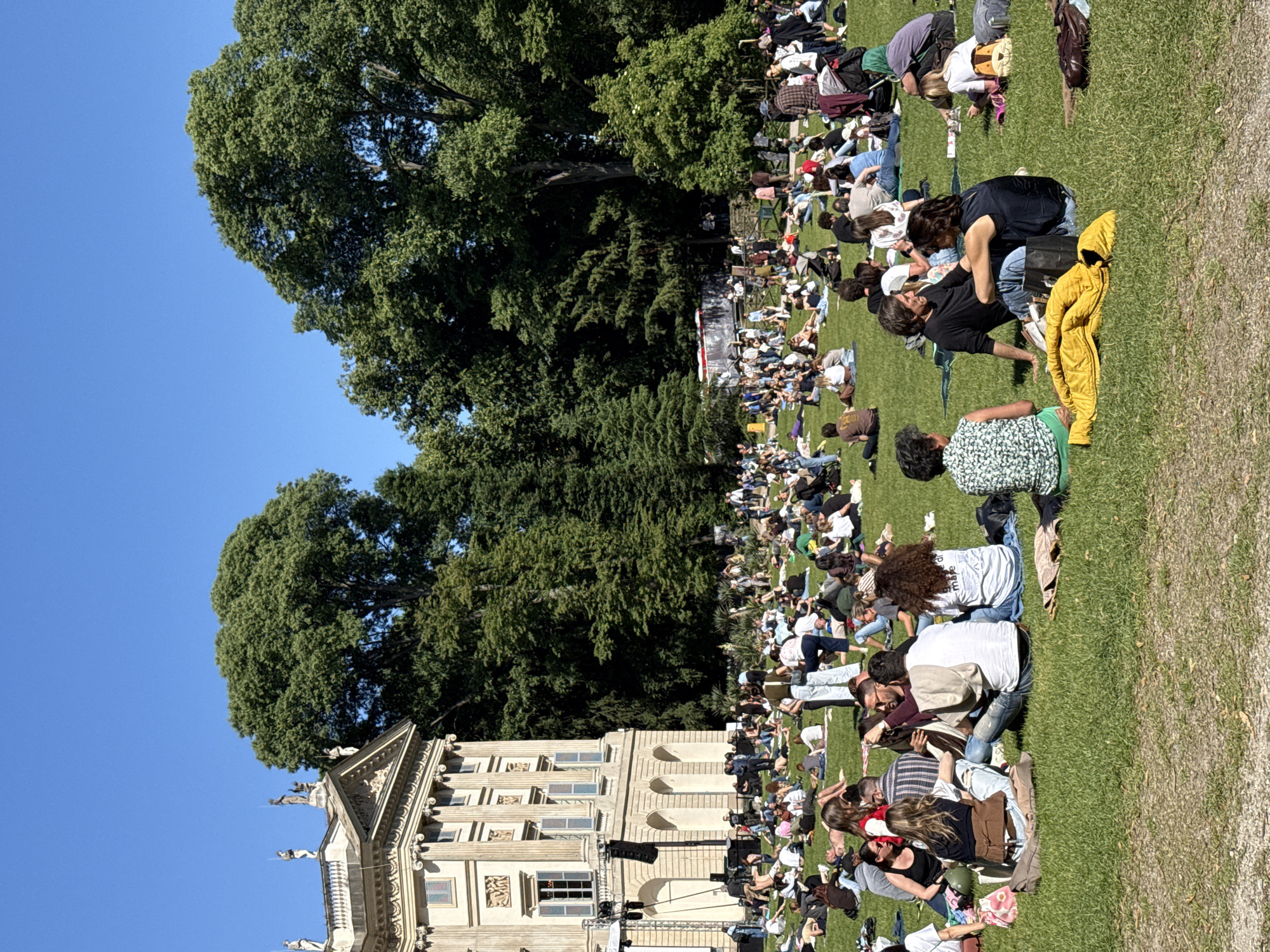 The image depicts a large crowd of people gathered on a grassy lawn in Milan, Italy. The people are seated or lying down, suggesting a relaxed atmosphere. In the background, a building with ornate architectural details is visible, along with large trees. The sky is clear and blue. The event appears to be a gathering or outdoor performance, given the stage-like setup near the building.