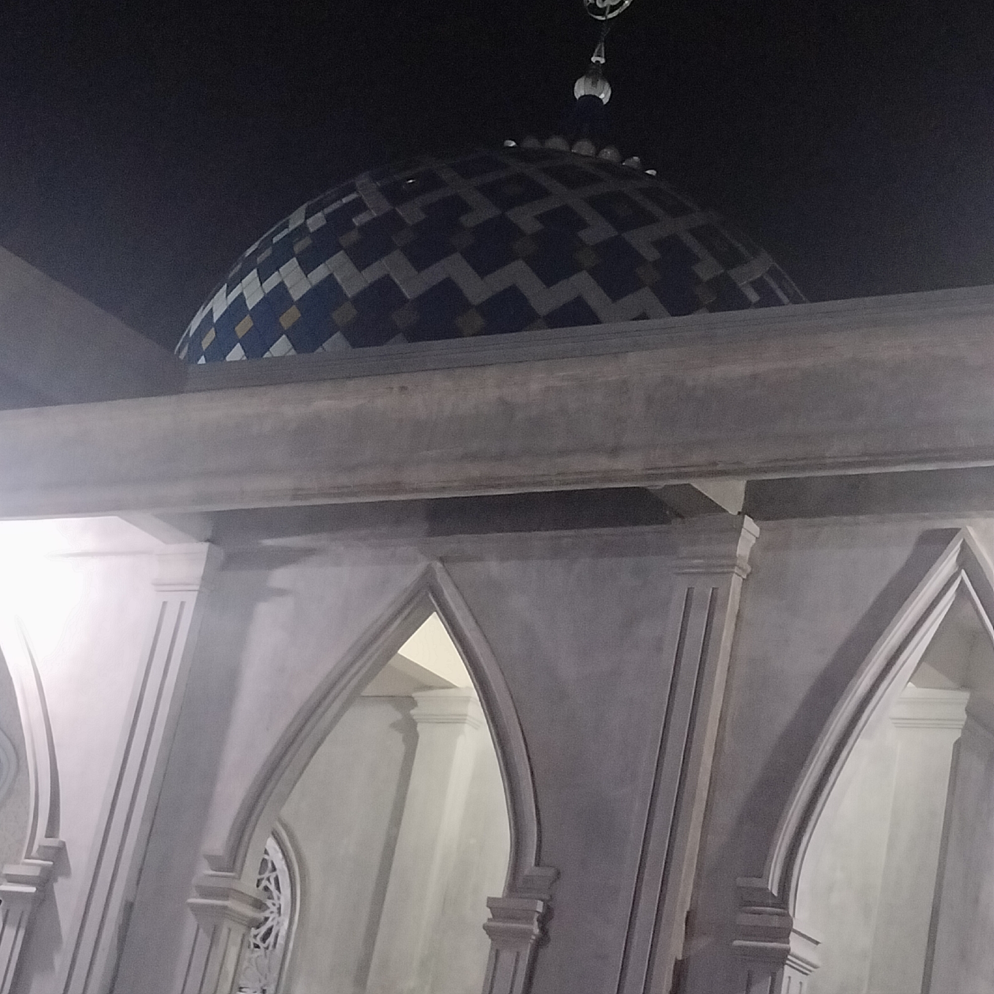 The image shows the architectural details of a mosque in Alueraya, Indonesia. Visible are the rounded arches, pillars, and a dome decorated with geometric patterns. The lighting is dim, possibly indicating nighttime. There are no people visible in the frame.