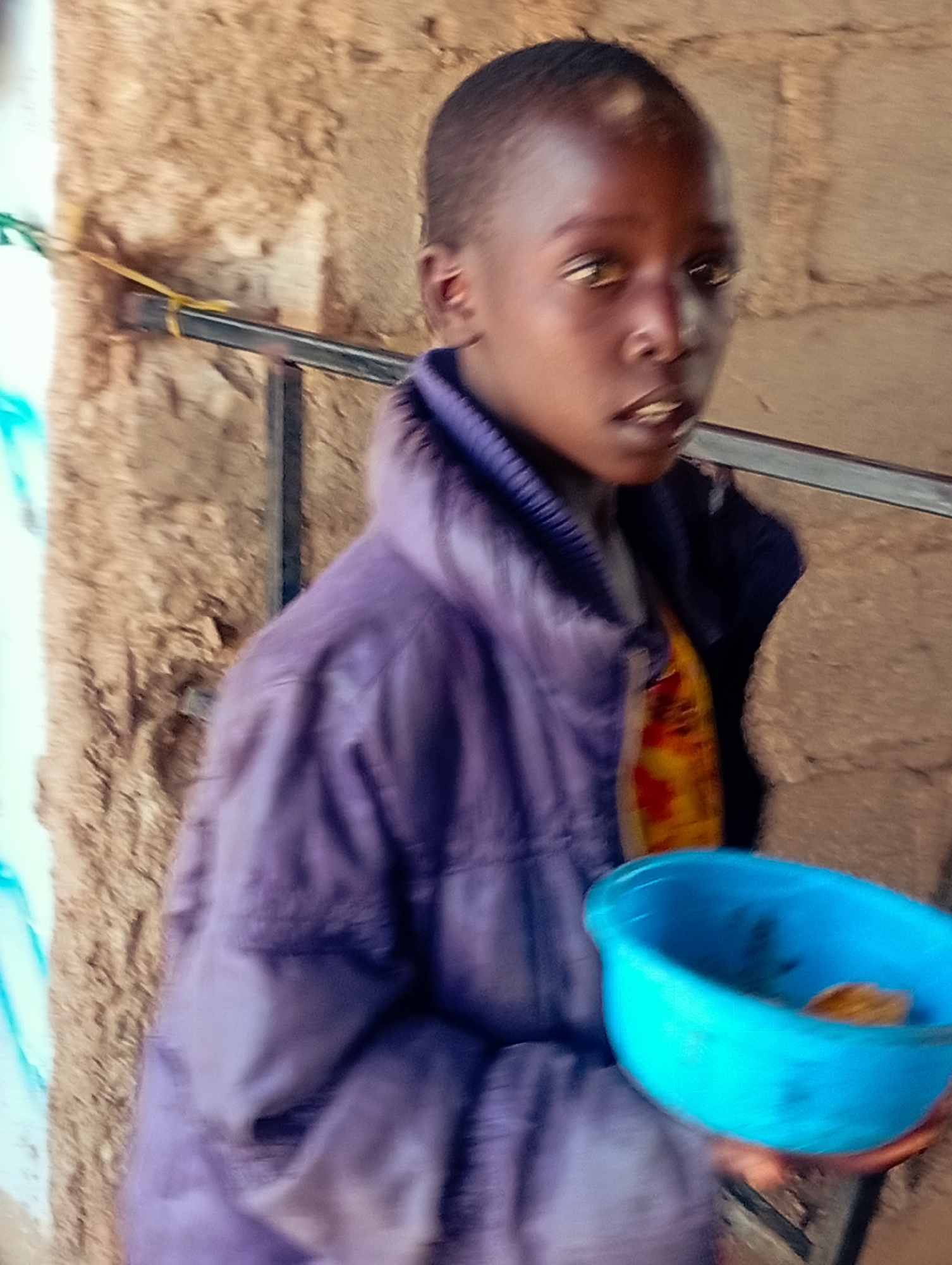 In Bukuru, Nigeria, a young person with dark skin and short hair is standing in front of a textured, light brown wall. They are wearing a purple, puffy jacket over a yellow and orange patterned garment. Their mouth is slightly open, and they are looking towards the right. In their hands, they are holding a bright blue bowl, which contains a small amount of brown food. A metal bar runs horizontally behind the person, and it is attached to the wall with a yellow cord on the left side. A blurry blue and white object is visible on the far left of the image. The lighting in the scene appears to be natural daylight.