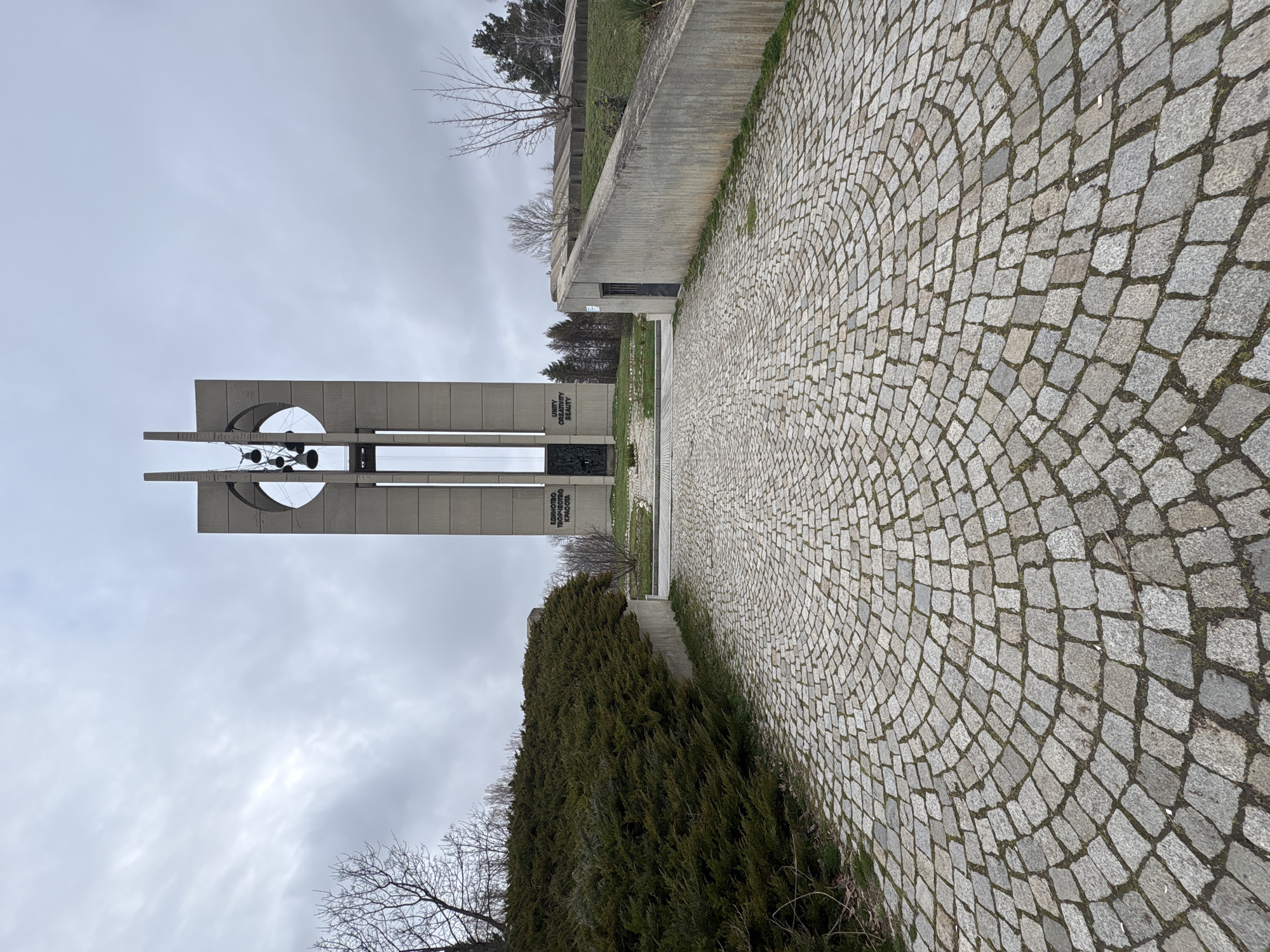 This image depicts the Bells Monument, located in Sofia, Bulgaria. The monument is constructed of several large concrete pillars, some of which are connected by a horizontal structure with a circular cutout. Metal bells are suspended within the cutout. To the right of the monument, a paved cobblestone walkway curves into the distance, bordered by a concrete wall and greenery. The sky is overcast. The monument has inscriptions in Bulgarian.