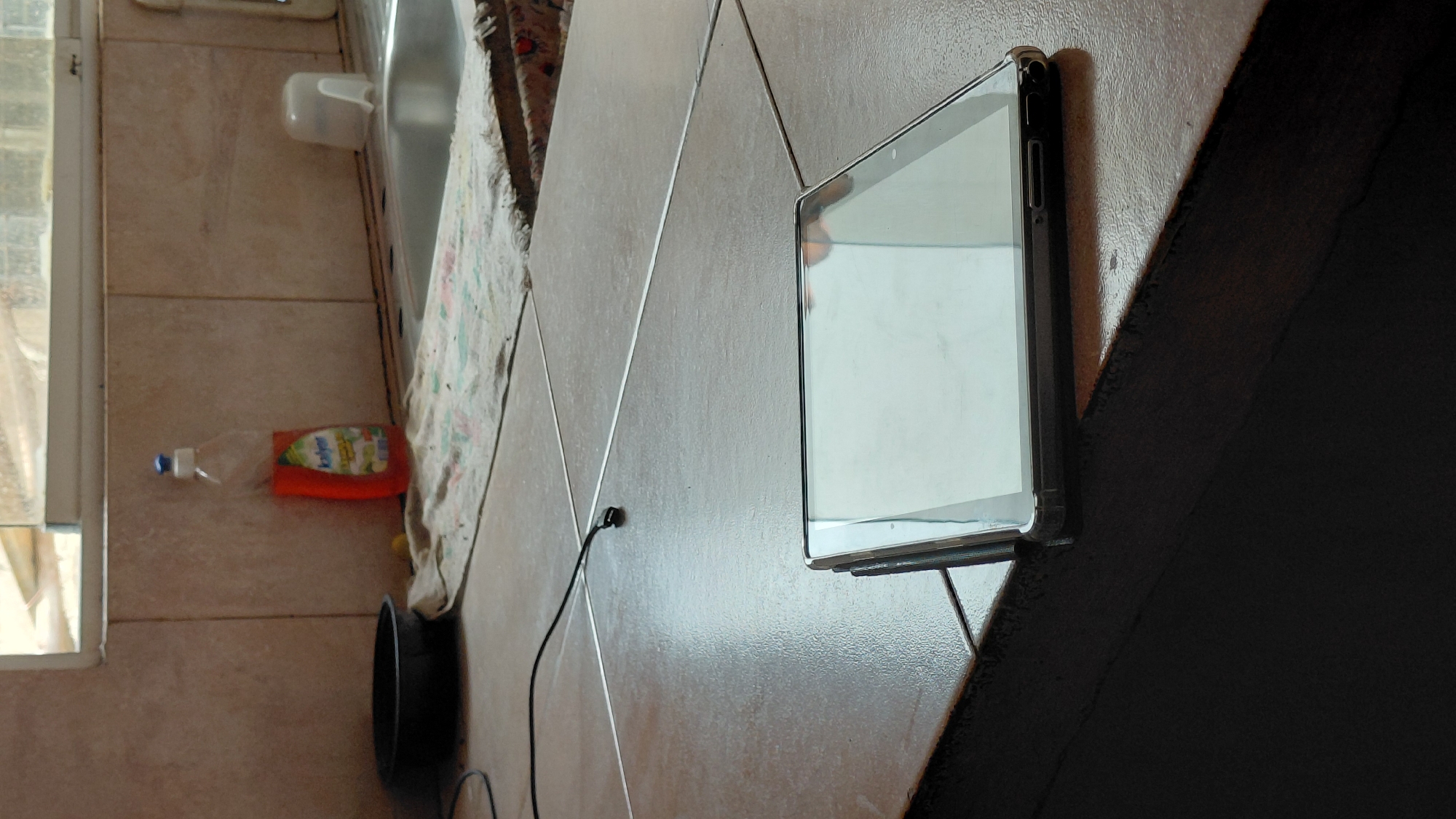 The image depicts an indoor scene, likely a kitchen counter, with a tablet device prominently displayed in the foreground. The tablet is silver and black, lying flat on a tiled surface. The tiles are brown and appear to be laid in a grid pattern, with thin metallic grout lines. In the background, a kitchen sink is visible, along with a bottle of orange dish soap and a white plastic cap. A dish towel lies near the sink, and a black circular object, possibly a bowl, is situated to the left. A black cable extends from the left side of the frame, disappearing under the tablet. The lighting suggests an indoor environment with natural light coming from a window above the sink. The image does not contain any explicit information indicating the city of Santa Ana, Venezuela.
