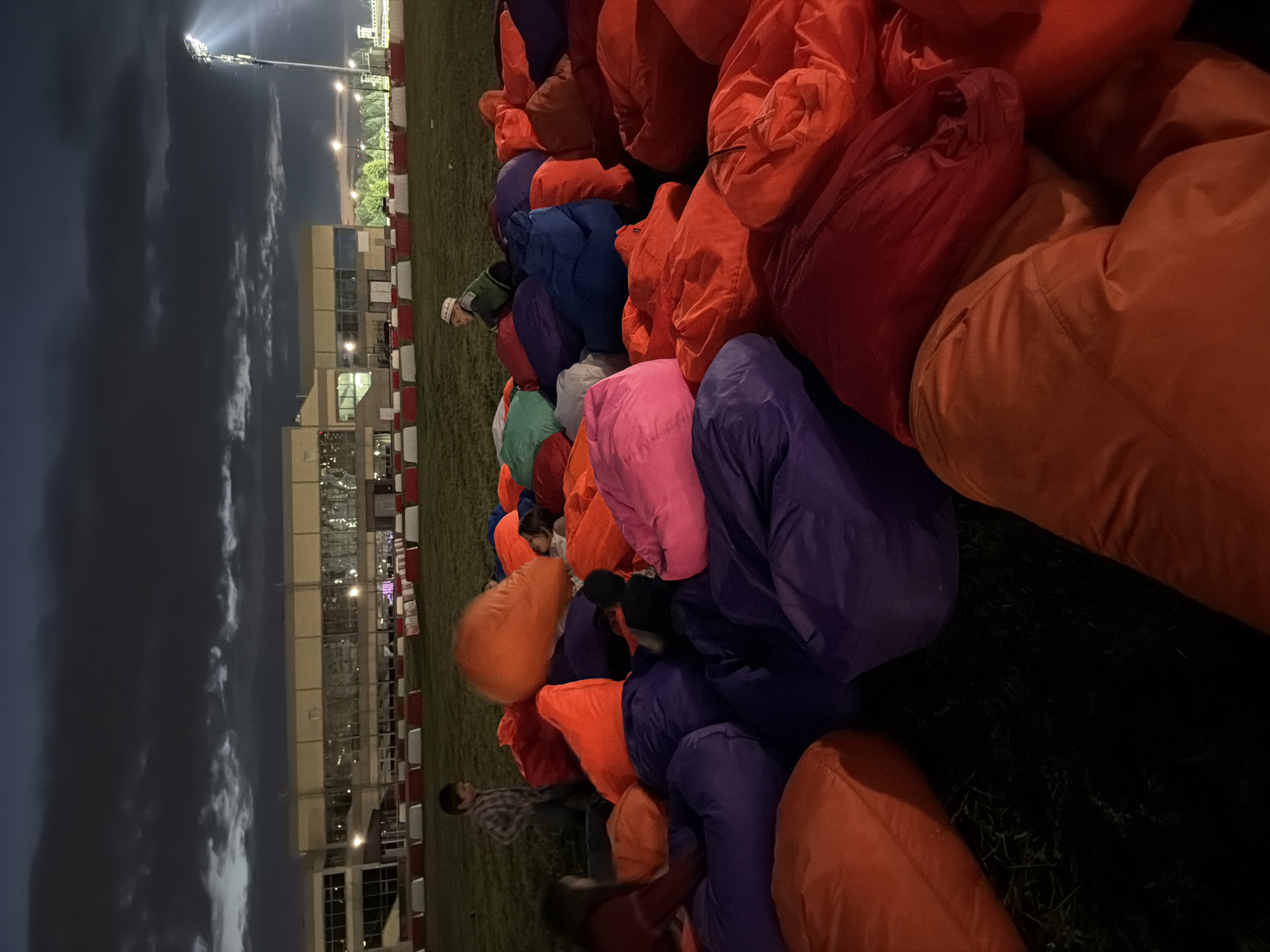 An outdoor scene at dusk features a large gathering of people on a grassy area covered with numerous colorful beanbag chairs. The beanbag chairs are predominantly orange, purple, and pink, creating a vibrant and informal seating arrangement. In the background, a modern building with large windows is illuminated, suggesting it might be a stadium or event center. A tall light pole with a bright spotlight shines from the right side of the frame, indicating artificial lighting for the event. The sky above is dark with clouds, typical of evening hours. Several individuals are visible amidst the beanbag chairs, some seated, others possibly interacting or moving around. One child in a plaid shirt is standing, looking towards the beanbag area. Another child wearing a white cap is sitting on a beanbag. The ground appears to be grass, with some visible blades and shadows. The overall impression is of a casual outdoor event, possibly a movie screening or concert, taking place at twilight.