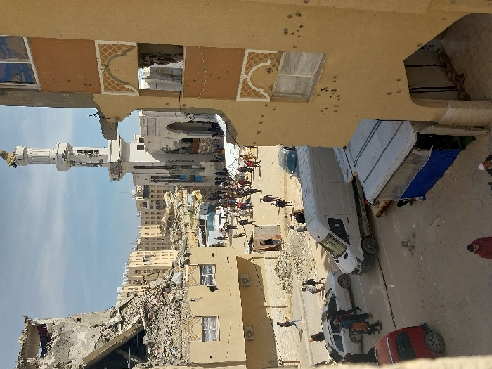 The image depicts a heavily damaged urban environment in Khan Yunis, Palestinian Territory, under a clear sky. In the foreground on the right, a multi-story, light-colored building exhibits numerous impact marks and apparent bullet holes across its facade. A large section of its lower wall is absent, forming an open archway, and a window on an upper floor is damaged. A temporary shelter with a light roof and blue side tarpaulin is situated at the base of this building.
The midground features a street with a mix of civilian pedestrians and vehicles. Approximately 50 to 100 individuals are visible, walking individually or in groups, with a larger crowd gathered further down the street. Vehicles include a white cargo van, a silver sedan, a dark sedan, and a red hatchback. The street and surrounding areas are strewn with rubble and debris from destroyed structures. On the left side of the street, two small, makeshift structures are present.
In the background, a white mosque with a tall minaret dominates the center, both showing visible structural damage. Surrounding the mosque and extending to the left are numerous multi-story buildings, many of which are severely damaged or partially collapsed, exposing interior elements and reinforcing bars. Extensive building rubble is distributed throughout the scene.