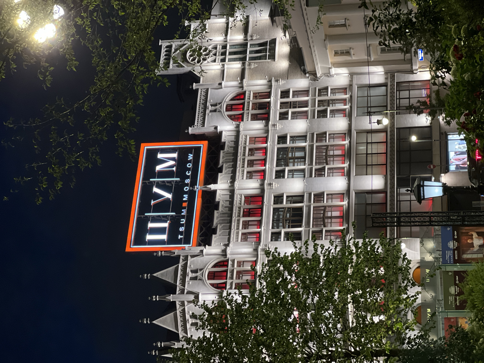 The image shows a nighttime exterior view of the TSUM department store in Moscow, Russia. The facade of the building is illuminated, with red lights visible behind many of the windows. A large neon sign for TSUM is prominently displayed at the top of the building, with the letters "ЦУМ" (TSUM) in Cyrillic, followed by "MOSCOW" in Latin script. Trees with green leaves are in the foreground, and a street lamp is visible on the right side of the image. The sky is dark.