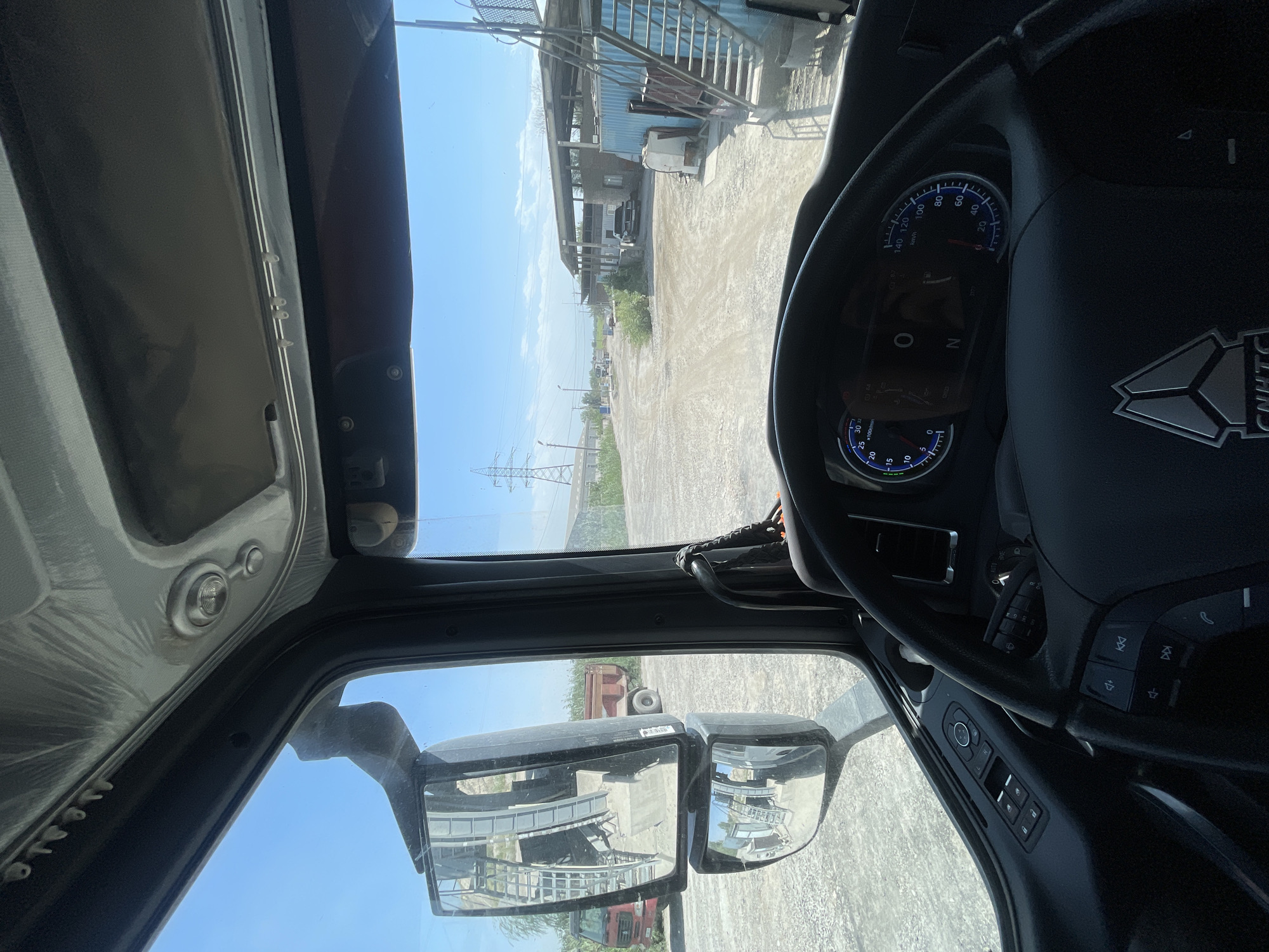 The image shows the interior of a truck's cabin, looking out through the windshield and side window. The steering wheel and dashboard are visible in the foreground. Outside, a dirt road leads through an industrial area with buildings, storage containers, and power lines under a clear blue sky. Two other trucks, one red and one with a red dump bed, are parked in the vicinity. The location is identified as Dmitrievka, Kyrgyzstan.