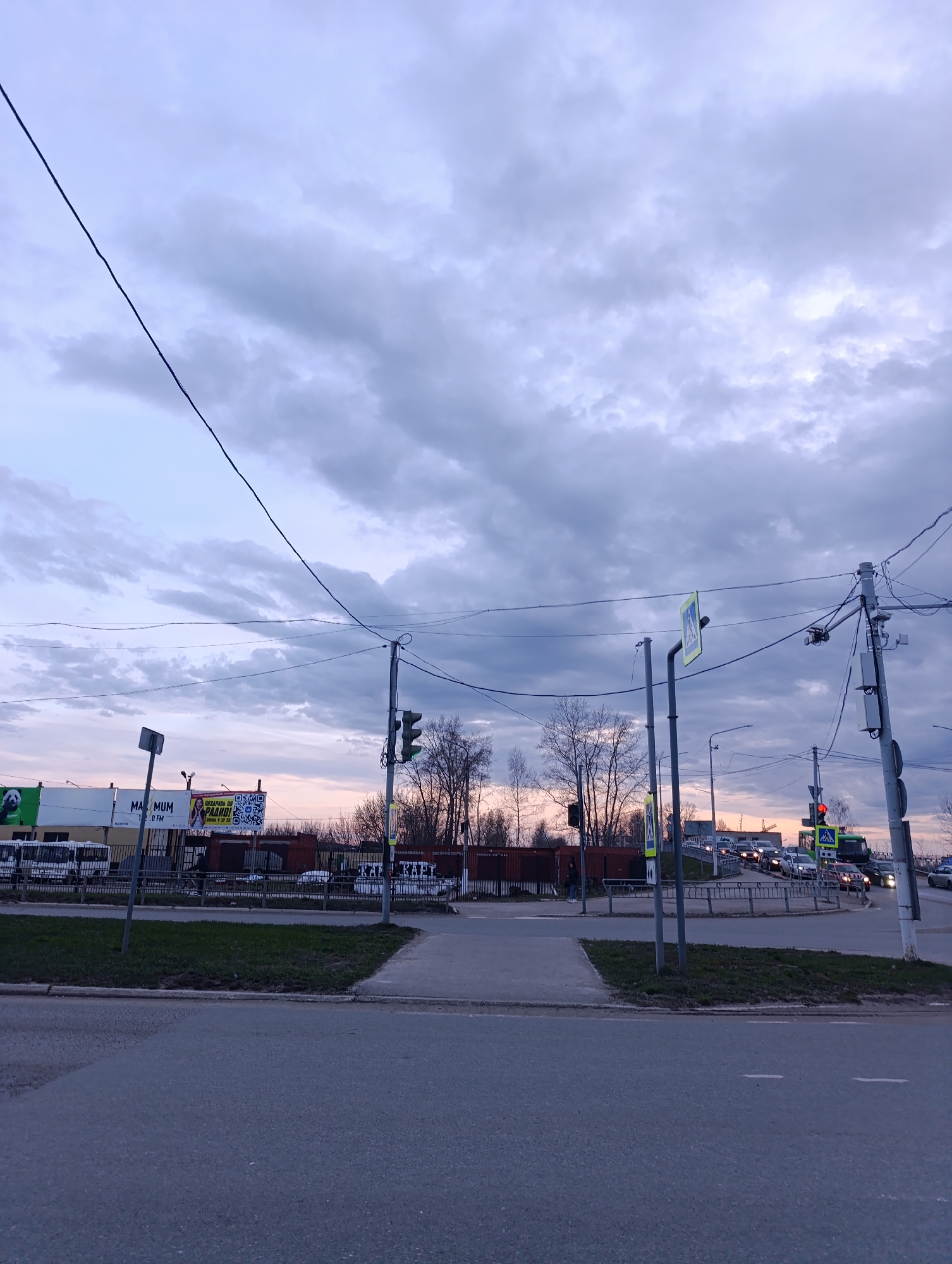 The image depicts a street scene in Chaikovsky, Russia. It is dusk or dawn, indicated by the colors in the sky. The location is a road with a pedestrian crossing. The crossing is marked with signs and traffic lights, which are red on one side. A group of cars is visible in the road, waiting for the light to change. There are some buildings on the side of the road, with commercial banners. There are overhead power lines. There is grass between the road and the sidewalk. There is one person visible, standing on the sidewalk.