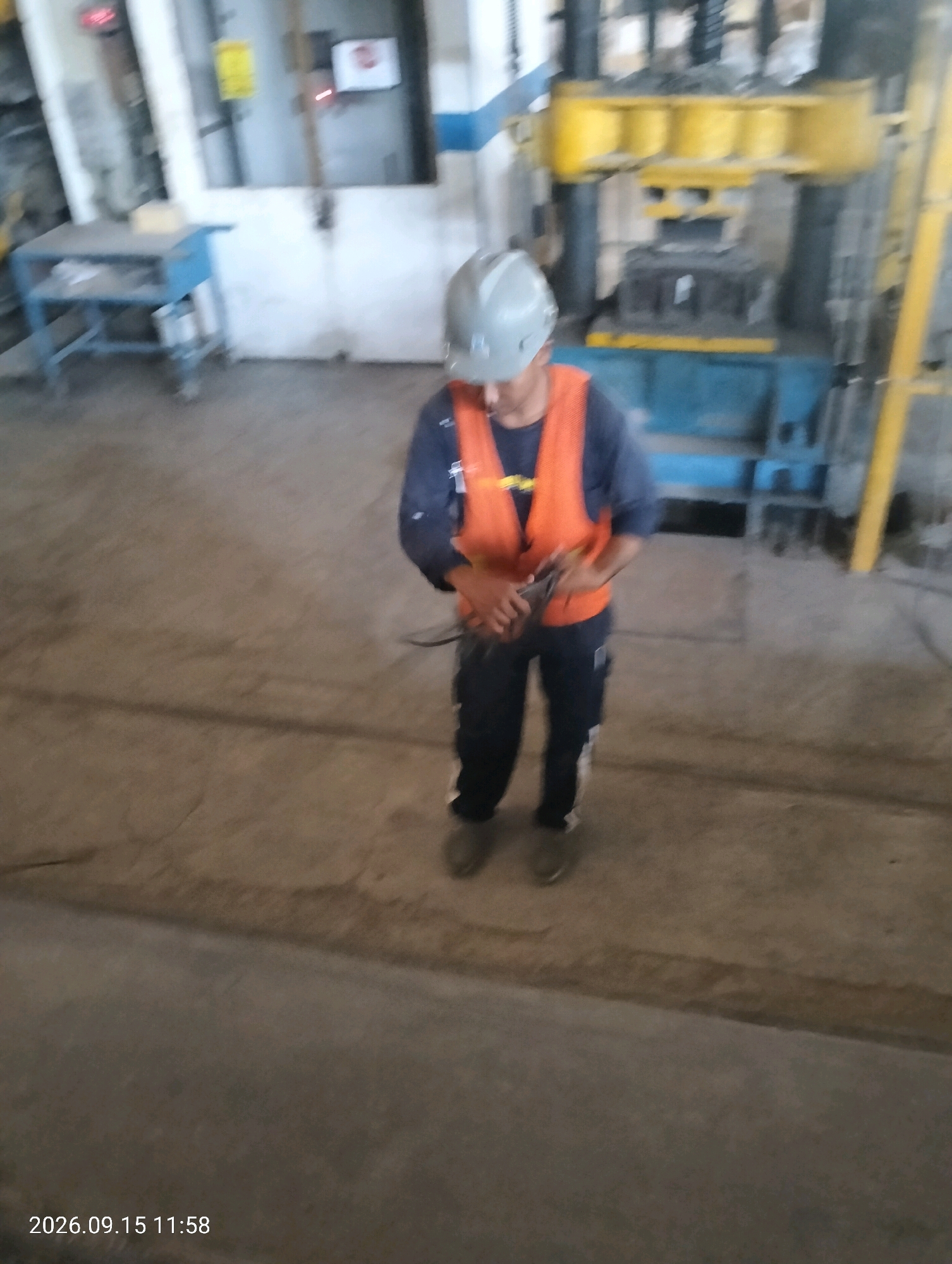 In an industrial setting, a person wearing a grey hard hat and an orange safety vest over a dark blue shirt is standing and holding a tool. The person is wearing dark pants and grey shoes. The background shows industrial machinery with yellow and blue components, and a blue table with shelves. The floor is concrete. A timestamp in the lower left corner reads "2026.09.15 11:58".