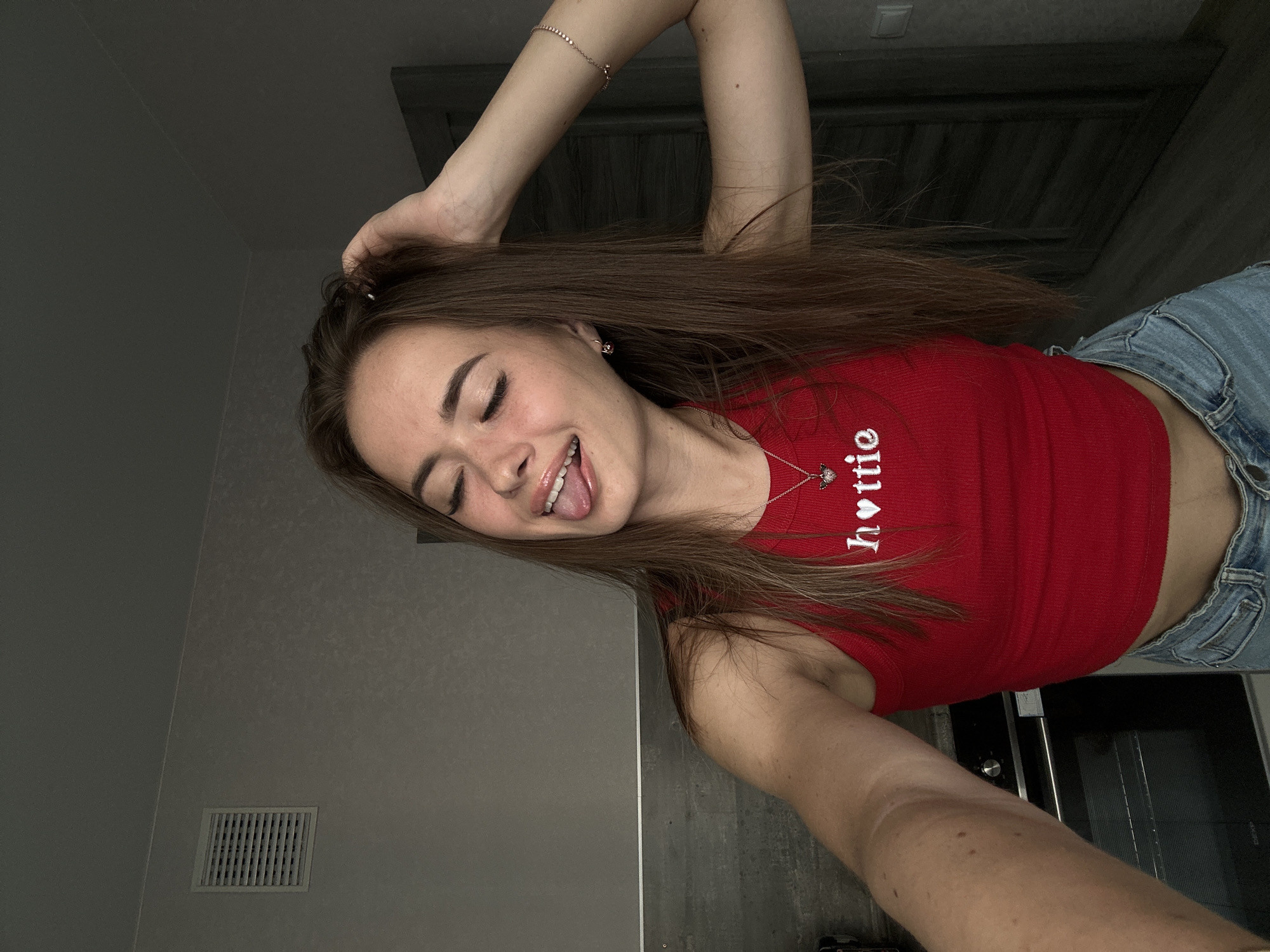 A young woman with long, brown hair and a red tank top with "hottie" written on it in white letters is posing for a selfie in what appears to be a kitchen. She is sticking her tongue out and closing her eyes, with her right hand in her hair. She is wearing denim shorts and a necklace with a heart pendant. The background includes a gray wall with a vent, a dark gray door, and a glimpse of a kitchen appliance.
