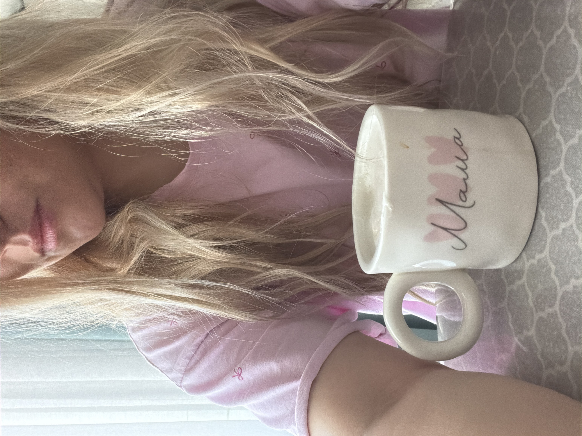 A woman with long blonde hair is lying down, wearing a pink shirt. Next to her on a patterned surface is a white mug with the word "Mama" written on it in a cursive script, accompanied by heart shapes. The location is not identifiable as Votkinsk, Russia.