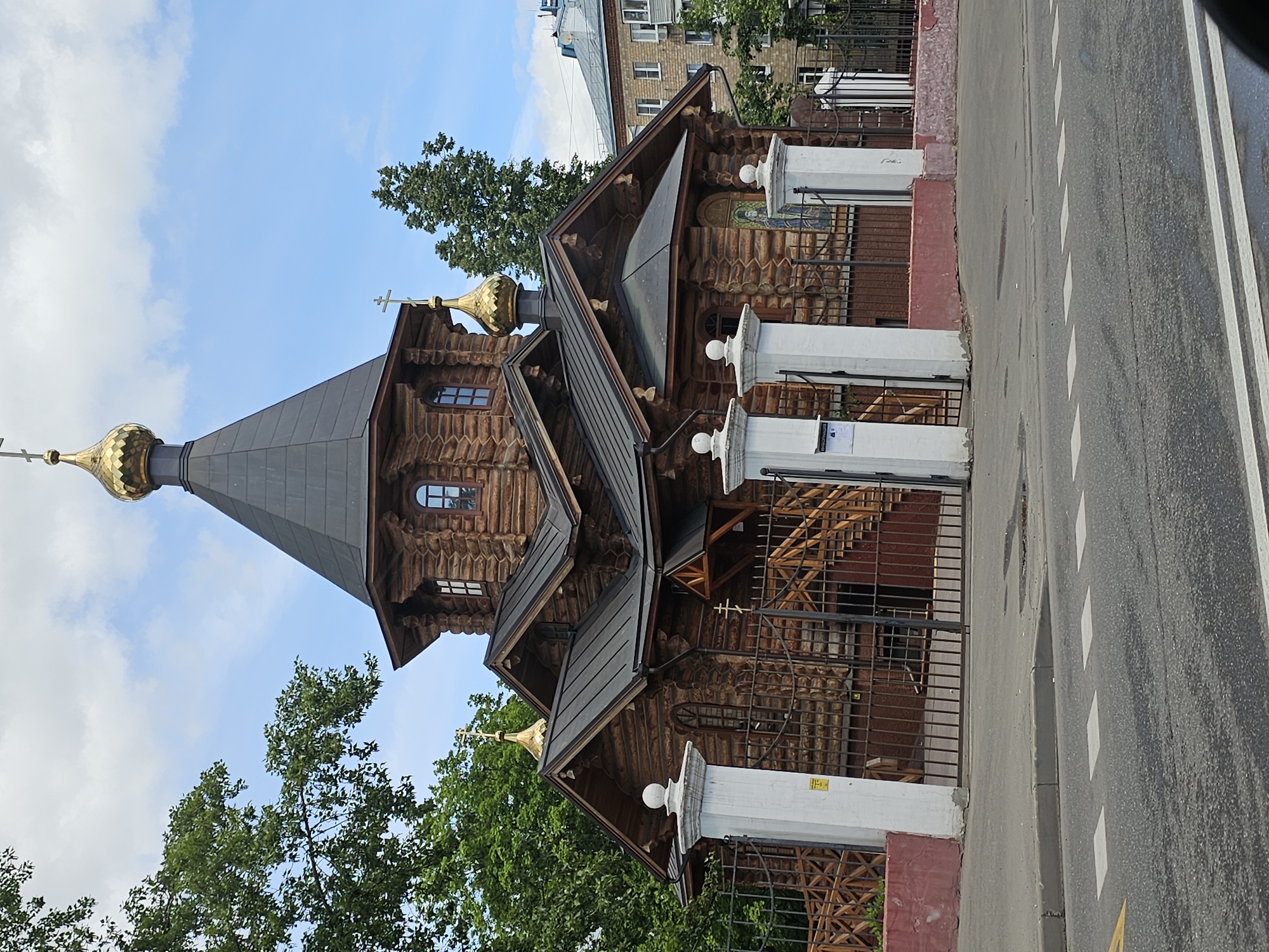 The image shows the Church of the Holy New Martyrs and Confessors of Russia, located in Khimki, Russia. The church is made of dark wood, with a gray conical spire topped with a gold dome and cross. The roof is black metal. The church is surrounded by a fence. A road runs along the side of the church. Trees are visible behind and to the sides of the church. The sky is blue with some clouds.