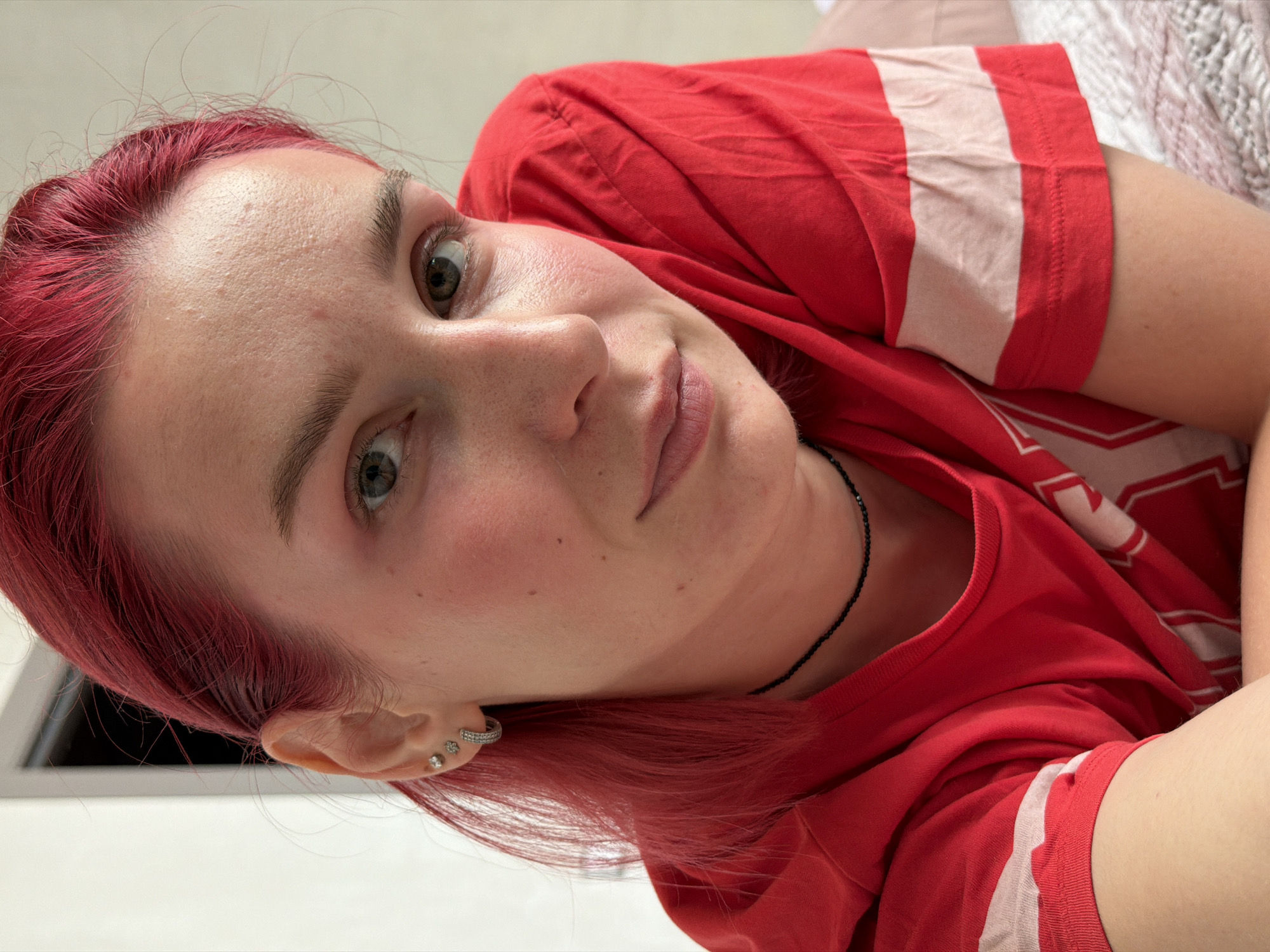 A close-up portrait of a young woman with vibrant red hair and light green eyes, looking towards the camera. She is wearing a red t-shirt with white stripes on the sleeves and a thin black choker necklace. The woman has several piercings on her left ear, including studs and a hoop. Her skin has a rosy hue, and there are visible pores and a few blemishes on her forehead and cheeks. The background is blurred, suggesting an indoor setting with a plain wall and a hint of what appears to be a bed with a textured blanket. The lighting is soft and natural. The image does not contain any specific elements that would allow for localization to Chaikovsky, Russia.