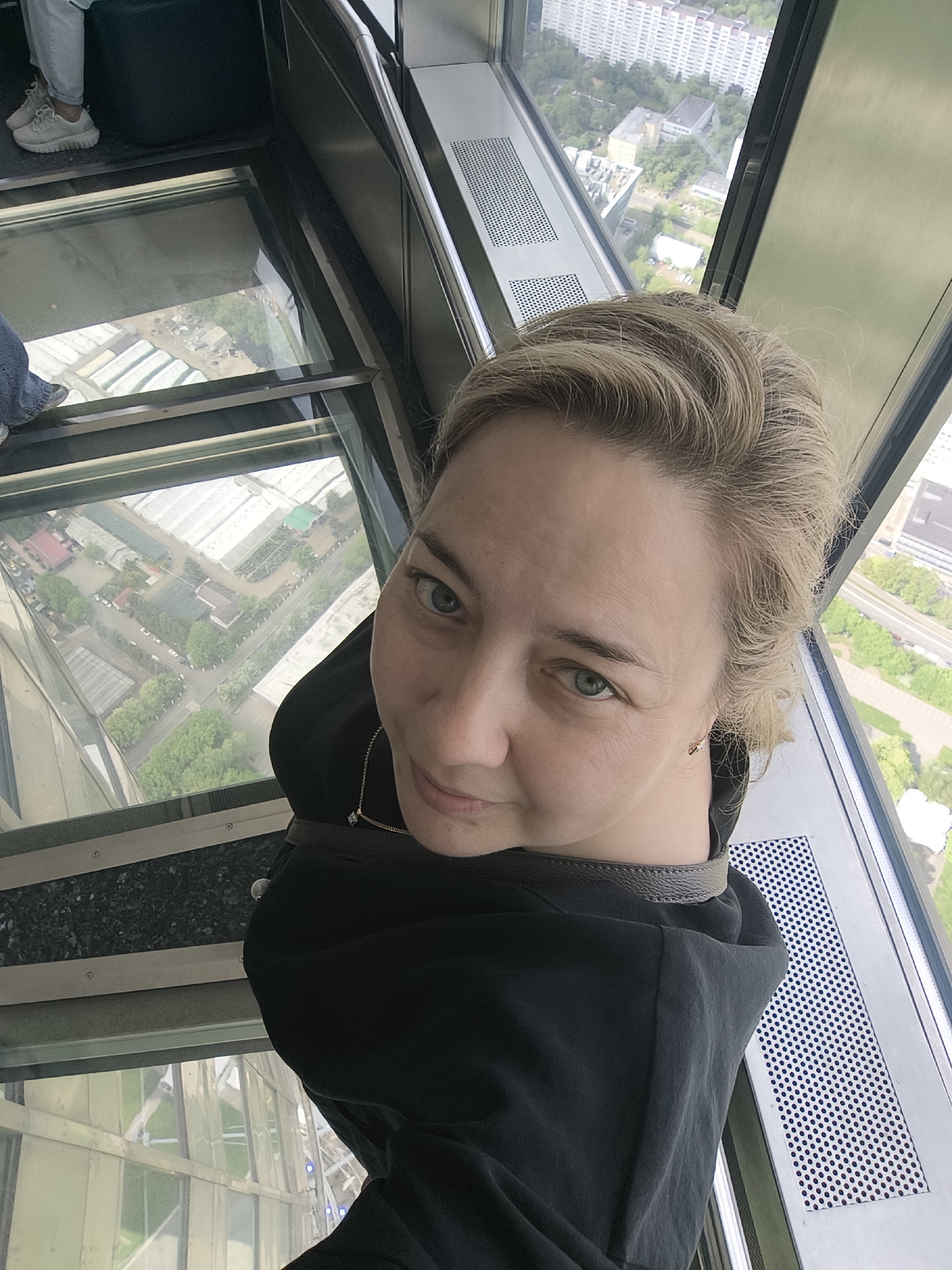 The image shows a woman taking a selfie inside a high-rise building with a glass floor. She is blonde and wearing a black sweatshirt. The glass floor allows a clear view of the city below, featuring buildings, streets, and green spaces. The location appears to be in a modern urban environment.