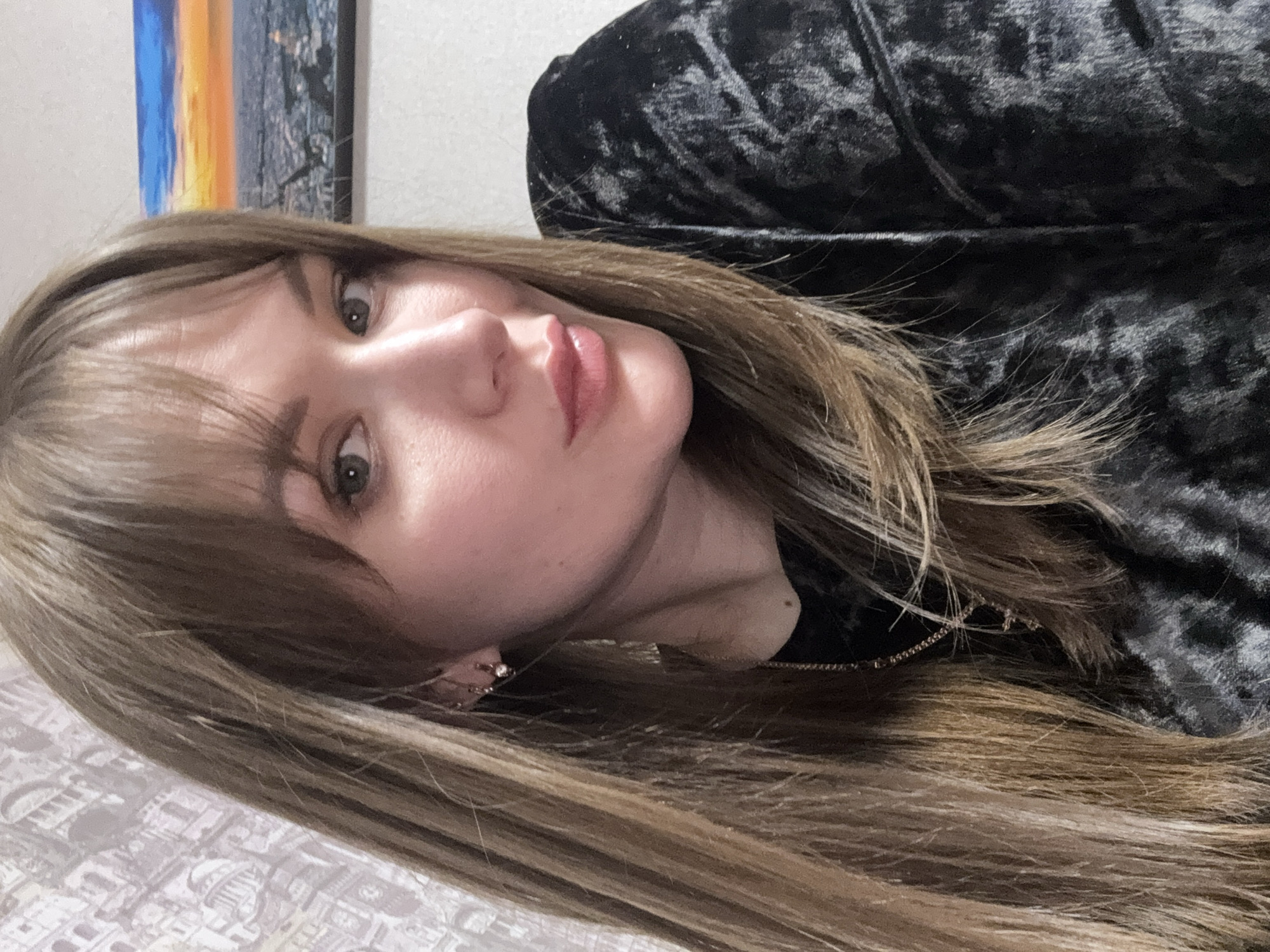 A medium shot depicts a Caucasian woman with light brown, shoulder-length hair and bangs. She has fair skin, light green eyes, and pink lips. She is wearing a dark, possibly velvet, top and a gold necklace. In her left ear, visible in the image, is a small earring.
Behind her is a blurred background featuring a patterned surface and an unfocused image, which appears to be a cityscape or nature scene with blue, orange, and white colors. The location of the image in Chaikovsky, Russia is not discernible from the provided image.