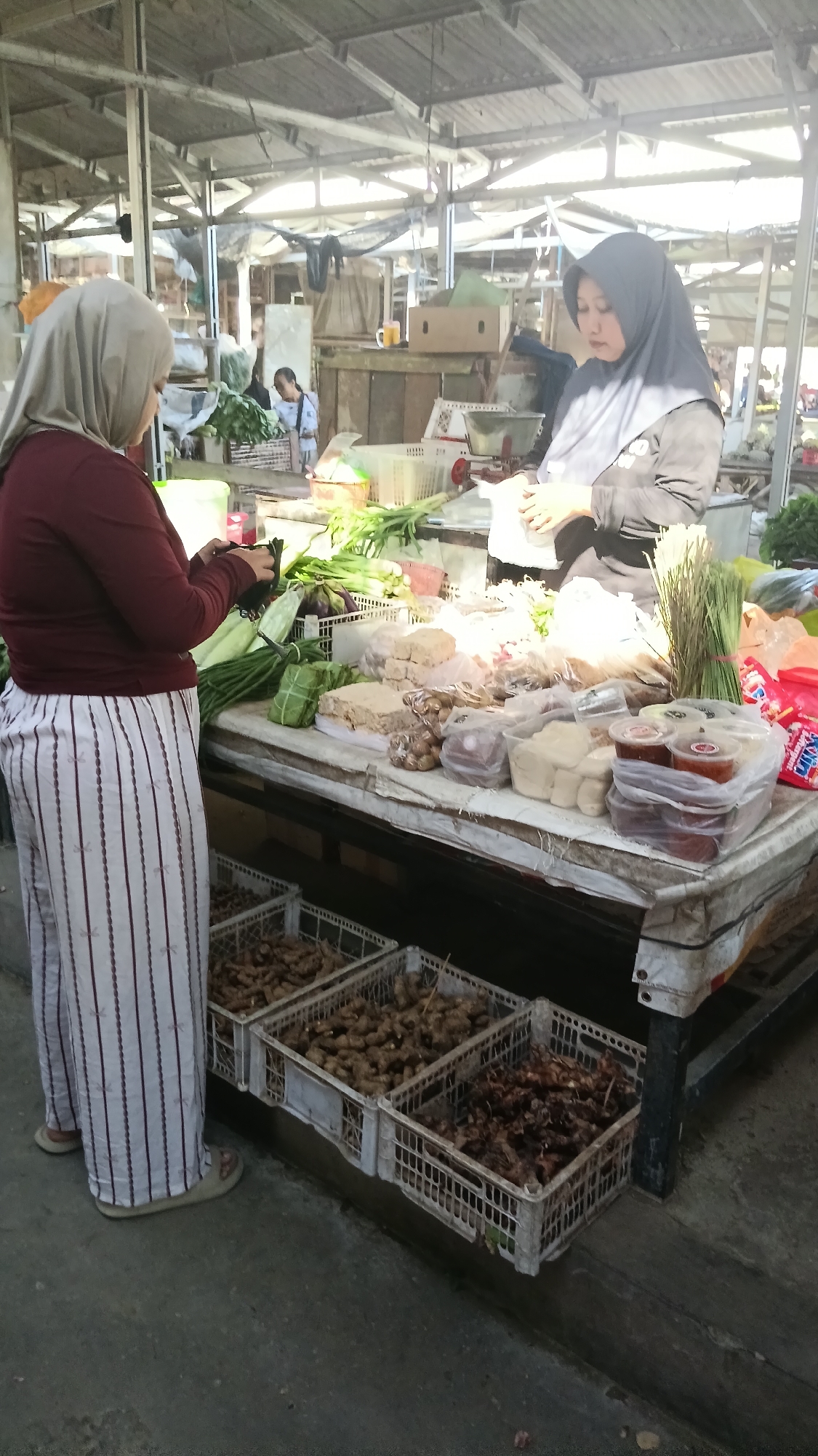 A woman in a light-colored hijab and a maroon long-sleeved shirt is standing on the left side of the image, facing a market stall. She is wearing striped white and maroon pants. She is holding a black object, possibly a phone, in her hands and looking down at it. A market stall, covered with a patterned tarp, is in the center and right of the image. A woman wearing a dark hijab and a black shirt is standing behind the stall, facing the first woman, and appears to be interacting with her. The stall is laden with various goods, including what appear to be bundles of green vegetables, white blocks of tofu, plastic containers of condiments, and bunches of herbs. In front of the stall, on the ground, are several white plastic crates filled with root vegetables, likely ginger or turmeric. The environment suggests an outdoor market, with a corrugated metal roof structure overhead, indicating it is likely in Dukuh, Indonesia, given the context provided. The lighting is bright, suggesting daytime.