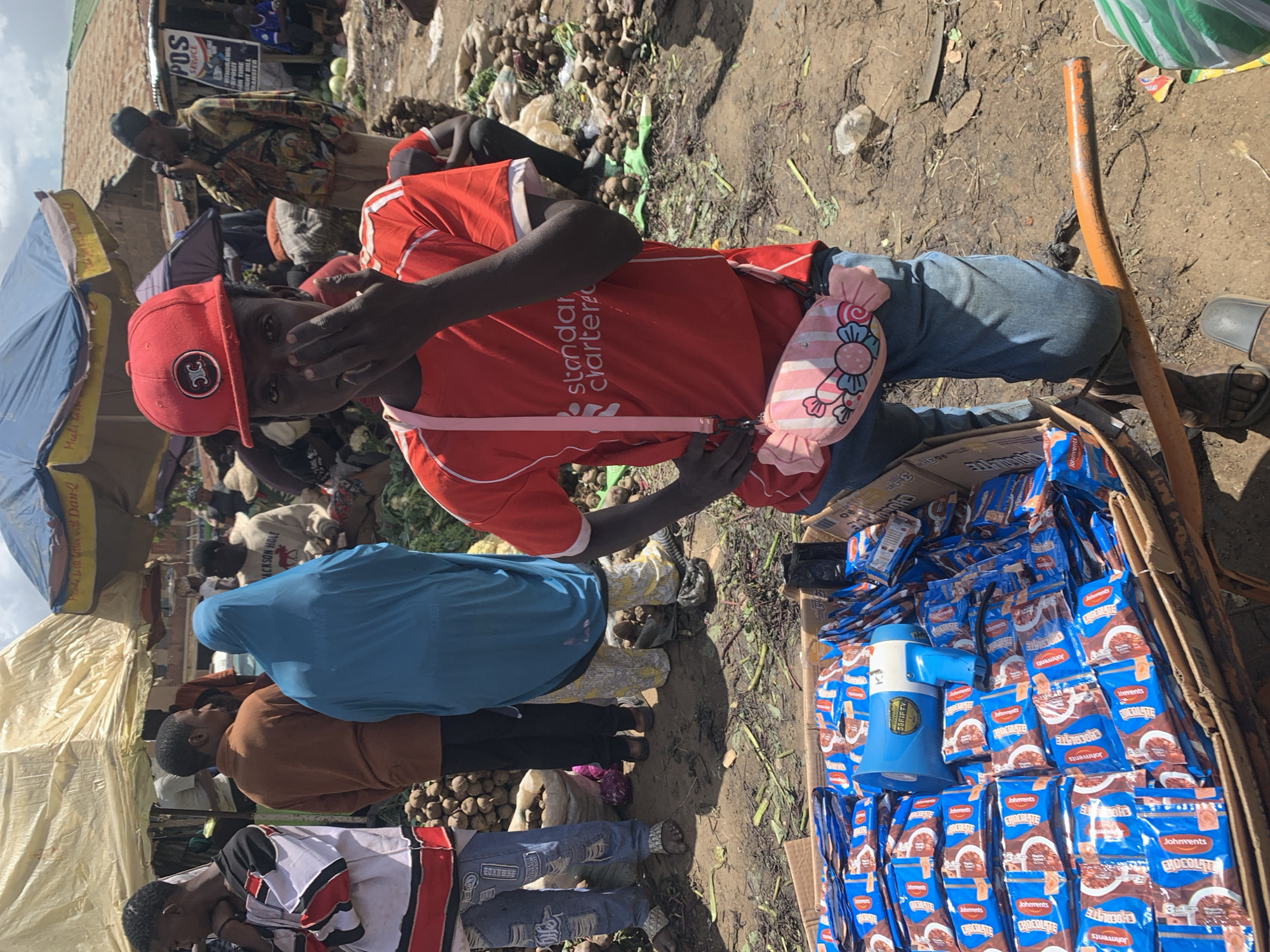 This media file depicts a lively outdoor market scene in Gura Top, Nigeria, under bright daylight. The ground is unpaved, dusty, and strewn with vegetable refuse and produce, suggesting an active, informal trading environment.
The central subject is a young person, possibly male, wearing a red baseball cap with a dark, stylized logo and a bright red t-shirt with "Standard Chartered" printed on the front. They are also wearing blue jeans and a distinctive pink, candy-shaped crossbody bag. Their left hand partially covers their face as they look towards the camera.
To the right of the central figure, a cardboard box is filled with numerous blue packages of "Johnvents CHOCOLATE" snacks. A blue and white megaphone or speaker, marked "59FIFTY," is placed among these items.
In the background, various other individuals are visible, including market-goers and vendors. Some are partially obscured by makeshift market stalls, which include tarps and blue and beige umbrellas providing shade. One blue umbrella has partially visible text "Prende taste better" and "Dan-". Another person wears a light blue head covering, and others are seen browsing or interacting. Piles of root vegetables, possibly potatoes, are visible on the ground in several areas. A "POS SERVICE" sign in the background lists services such as "WITHDRAWAL," "DEPOSIT," "TIME," "SIGHT BILL," and "TRANSFER." The overall activity indicates a bustling day of commerce and community interaction.