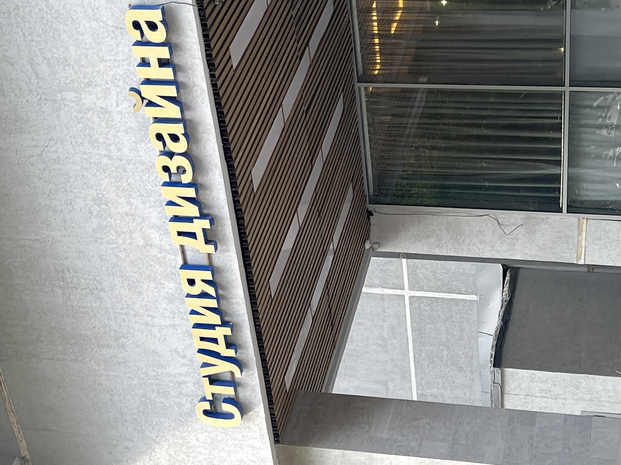 The image shows a close-up of a building facade with a sign that reads "СТУДИЯ ДИЗАЙНА" in large, yellow, three-dimensional letters with blue trim. The sign is mounted above a covered walkway with a slatted wooden ceiling and recessed lighting. To the left of the sign, a concrete column supports the roof structure. The wall behind the sign is textured and light gray. Below the covered walkway, large windows with dark curtains are visible, reflecting some of the interior lighting. The overall scene suggests an entrance to a commercial establishment in Moscow, Russia.