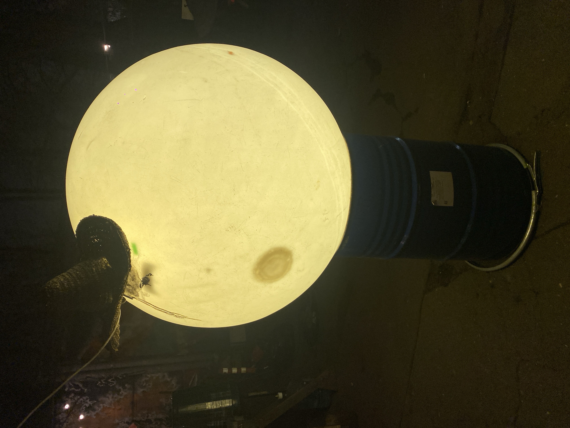 A street-level, nighttime view presents a large, illuminated, yellowish-white sphere connected to a dark, cylindrical barrel. The sphere is positioned on the left side of the barrel. A piece of dark, possibly woven material is attached to the sphere's surface on the left side. The barrel is secured with a metallic clamp. The ground appears to be paved or made of asphalt. The location is Moscow, Russia, but the precise street or landmark cannot be identified from the image alone.