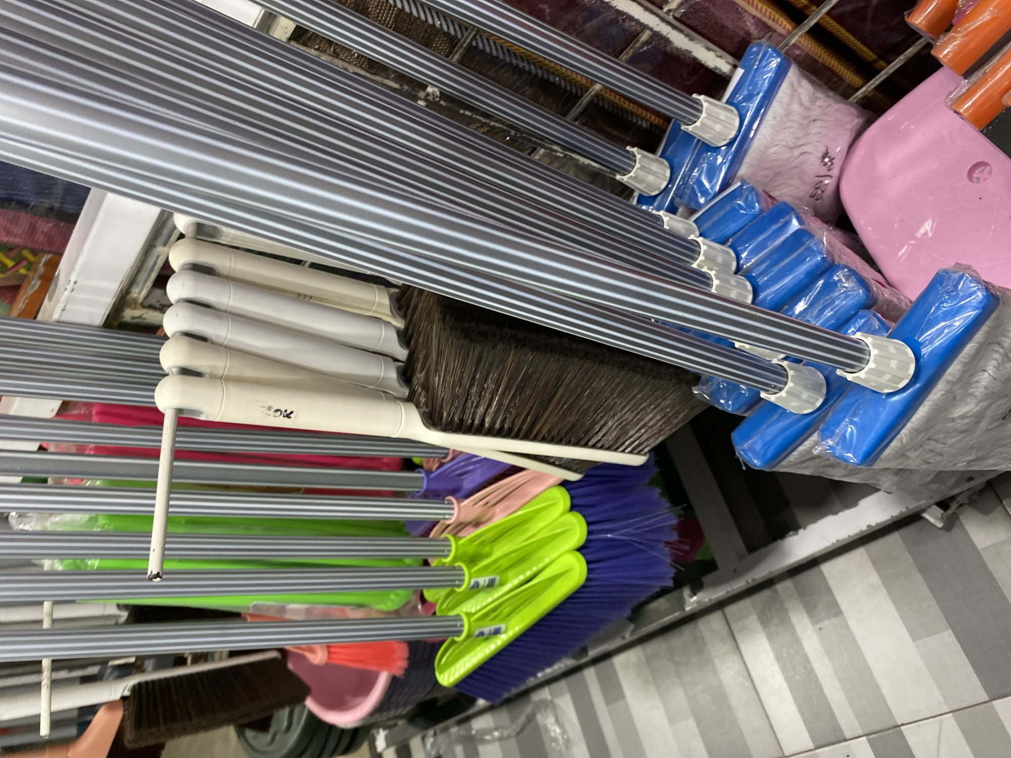The image displays a retail display of cleaning supplies, specifically brooms and mops, likely in a shop in Lamteumen Barat, Indonesia. A variety of brooms with colorful plastic handles and bristles in shades of green, purple, and orange are visible. Some brooms have thin, silver-striped metal handles, while others have white or cream-colored handles. Several mops with blue plastic bases and white fabric heads are also present, individually wrapped in plastic. A bright pink plastic object, possibly a dustpan or bucket, is situated at the bottom right of the frame. The background shows shelving units and other merchandise, suggesting a commercial setting. The floor appears to be tiled with a pattern of light and dark gray squares. No people are visible in the image.